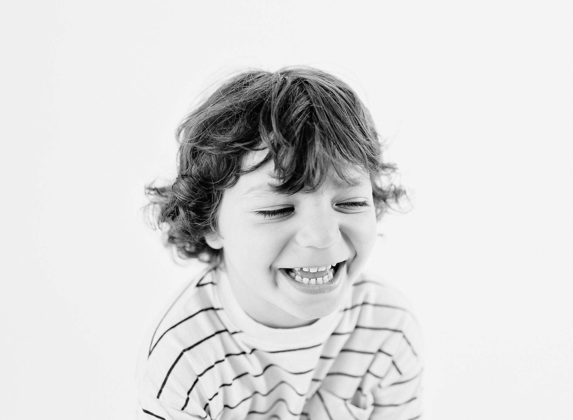 Boy with curly hair laughing, wearing a striped shirt, against a white background.