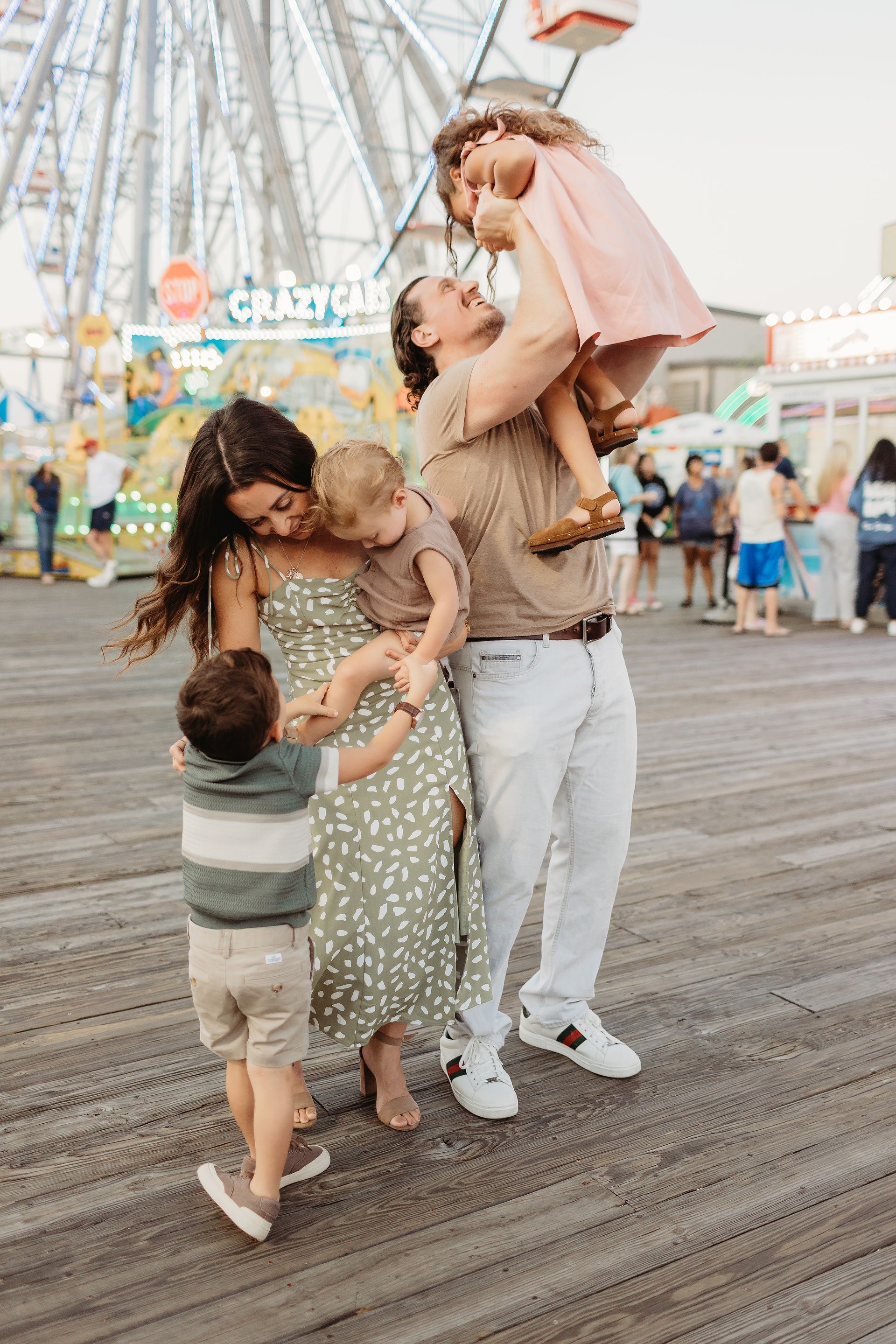 Family of five smiles at a boardwalk; father lifts a daughter.