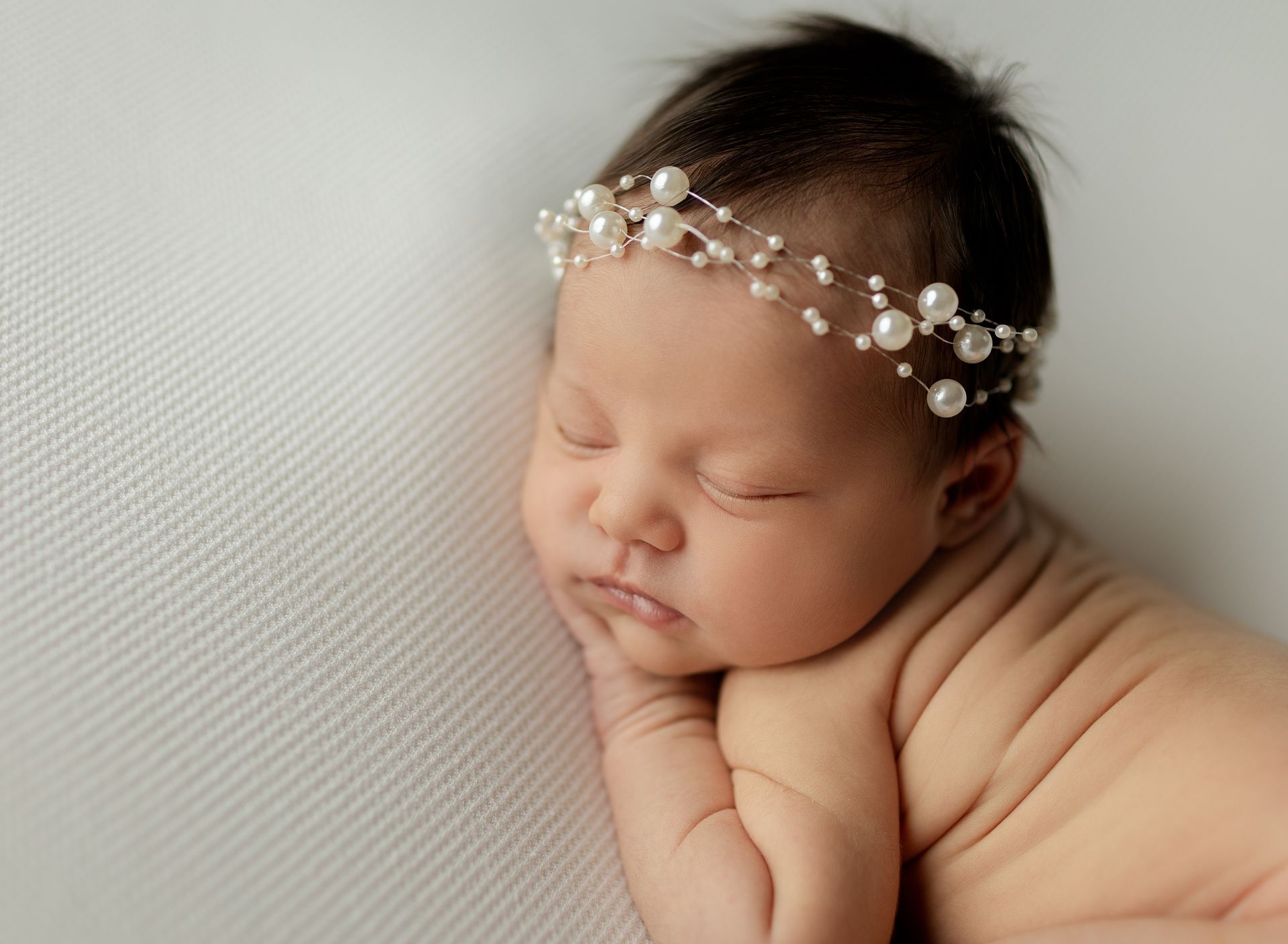 Newborn baby sleeping, wearing pearl headband, on white textured fabric.