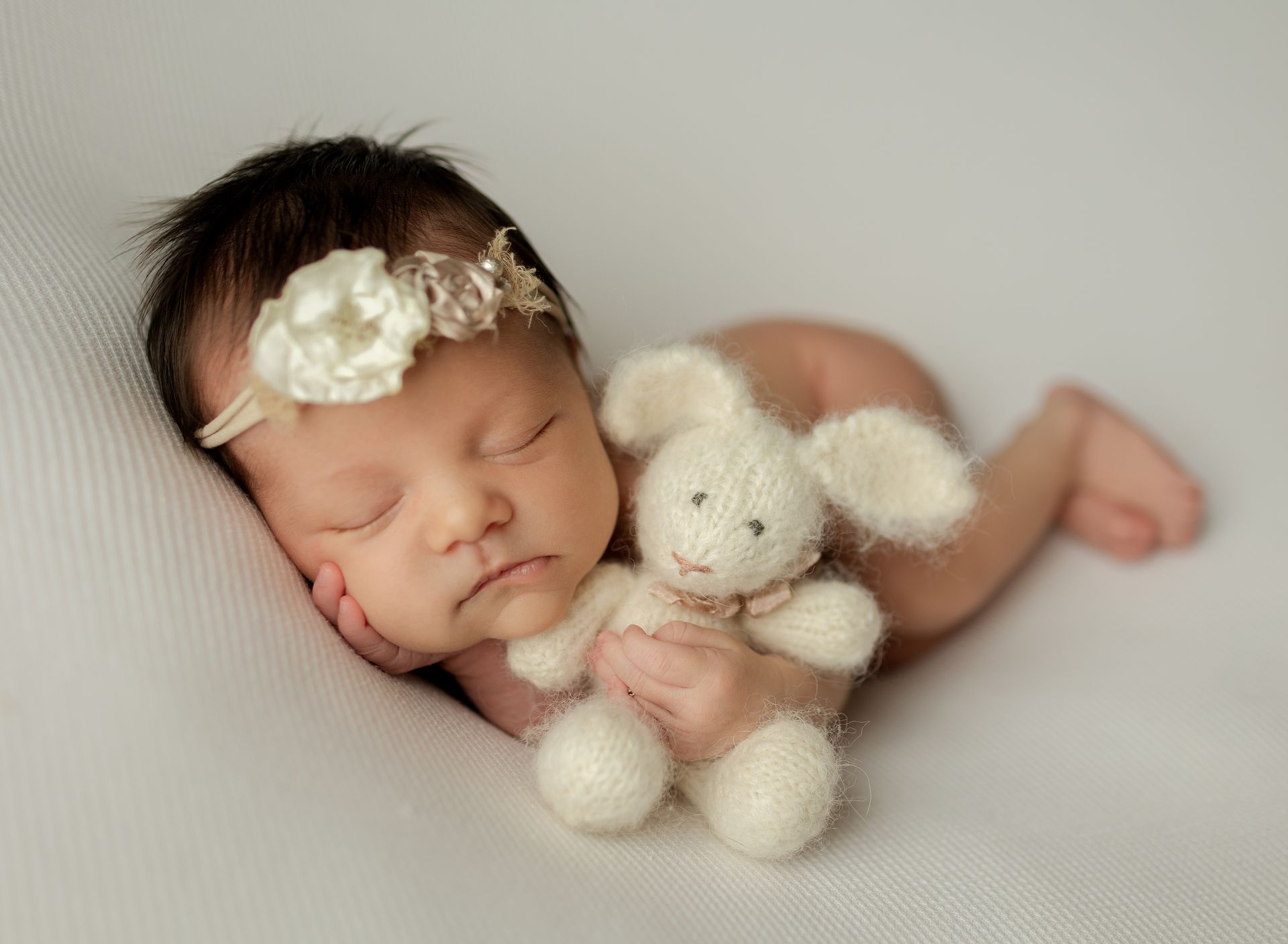 A newborn baby girl is sleeping on a white blanket holding a stuffed rabbit.