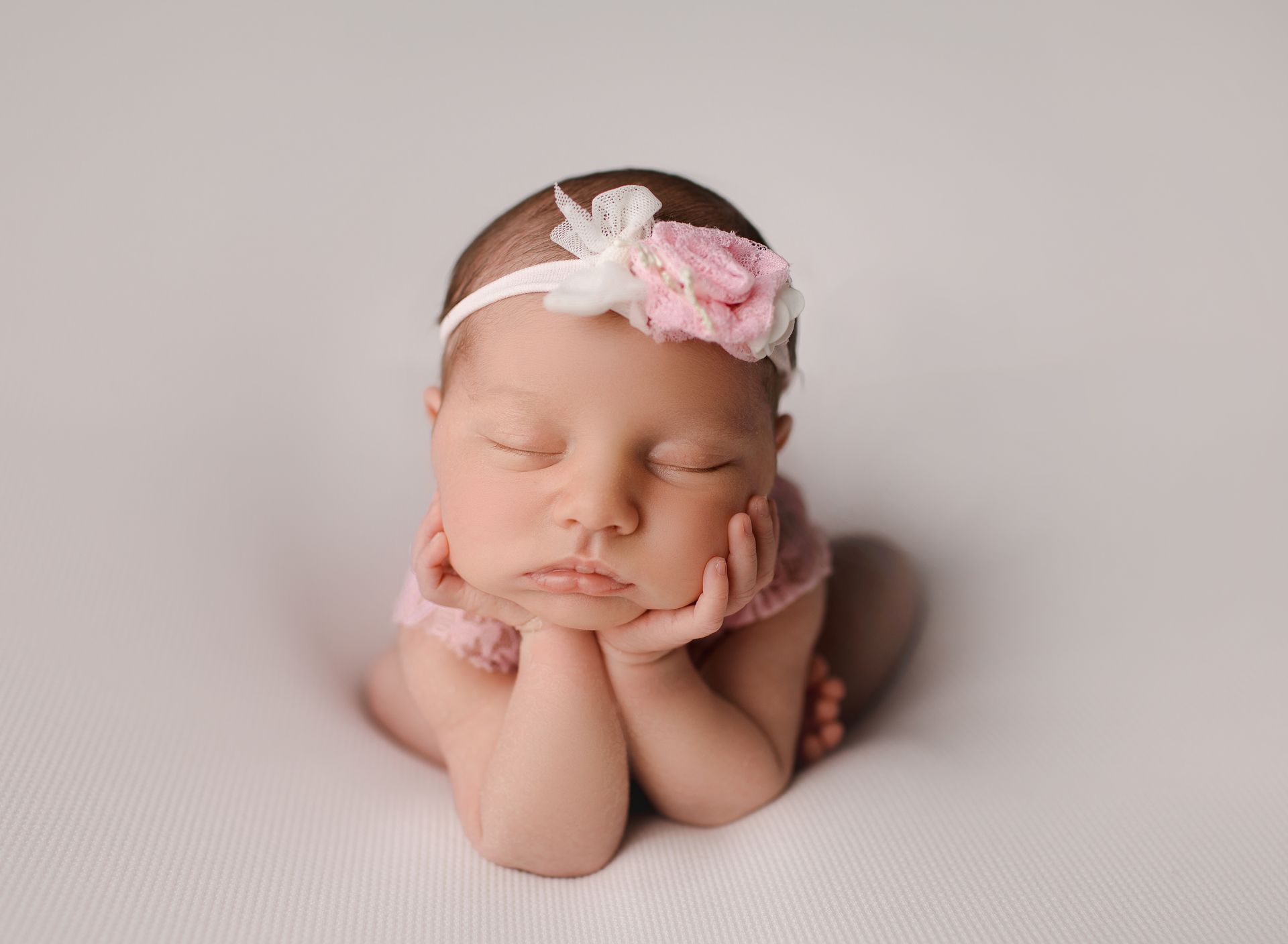 A newborn baby girl wearing a pink headband is sleeping on a white blanket.