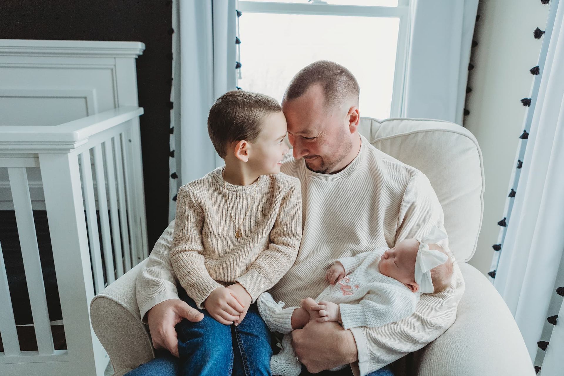 A father in a textured cream sweater sits in a chair, holding a baby while a child sits on their lap, looking at them in Glen Rock, NJ.