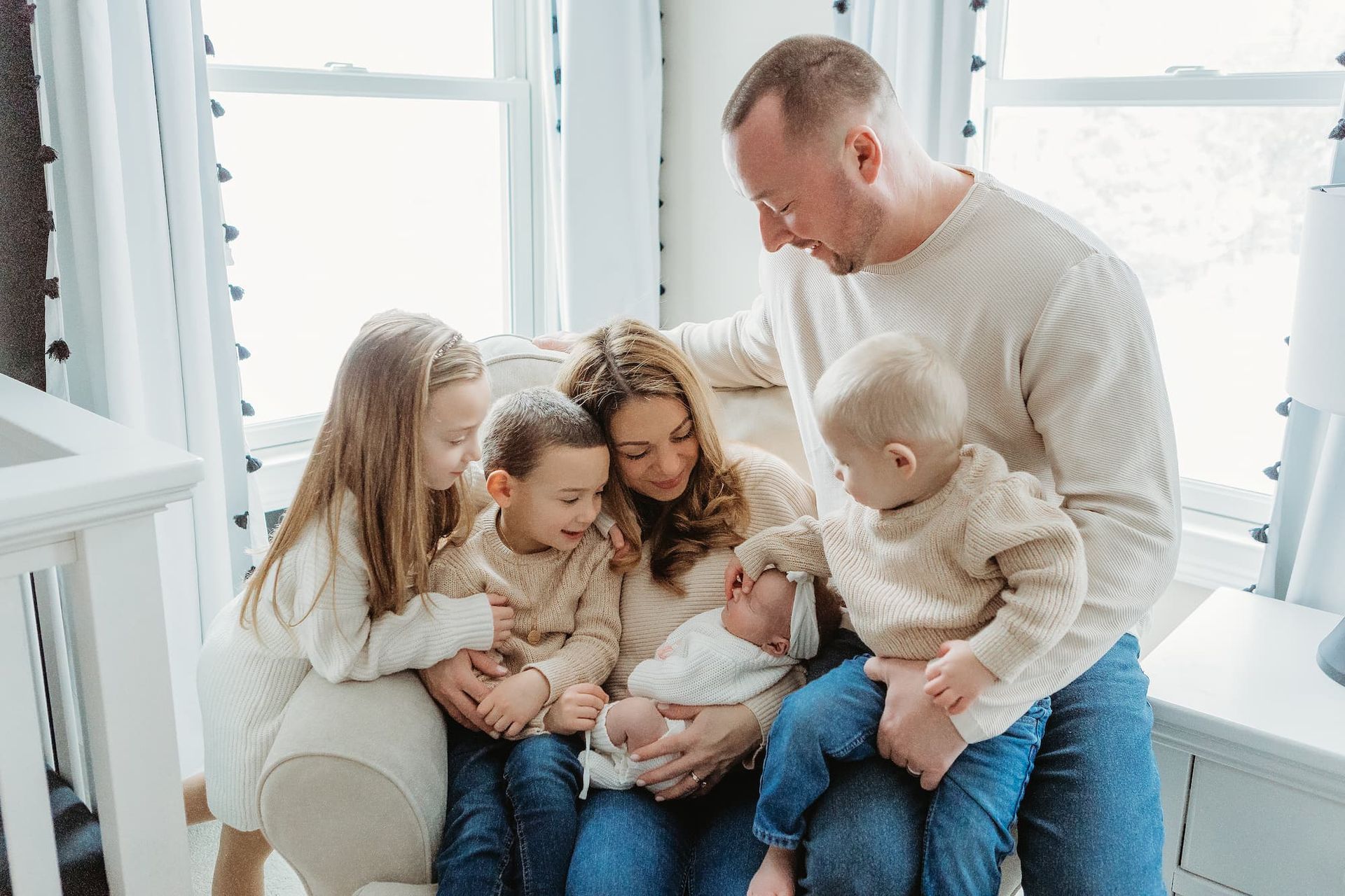 A family sitting together in a sunlit room, with parents holding a newborn baby surrounded by three children.