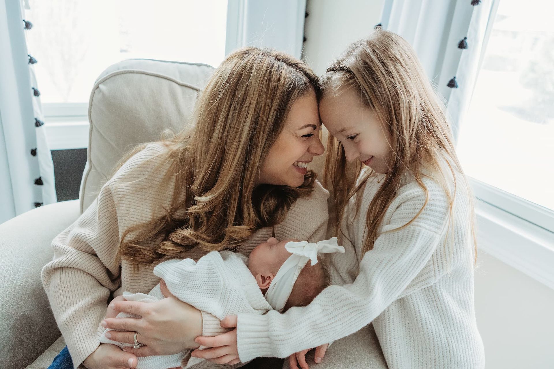 A mother holding a swaddled infant while a child leans in with their heads touching, all smiling in a light-filled room.
