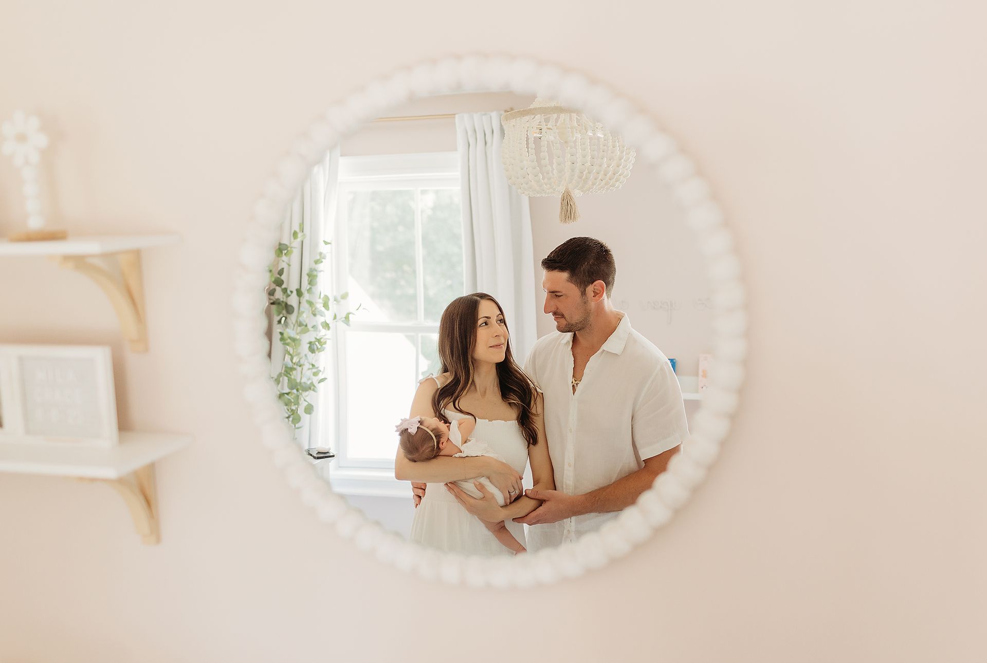 Family in mirror: Parents holding newborn, wearing white. Pink room, white beaded mirror frame in Passaic County, NJ