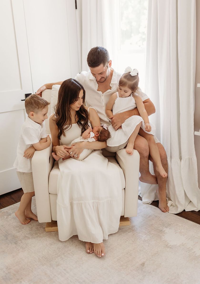 Family with newborn sits in a light-filled room; parents and children are wearing white clothes in Bergen County, NJ.