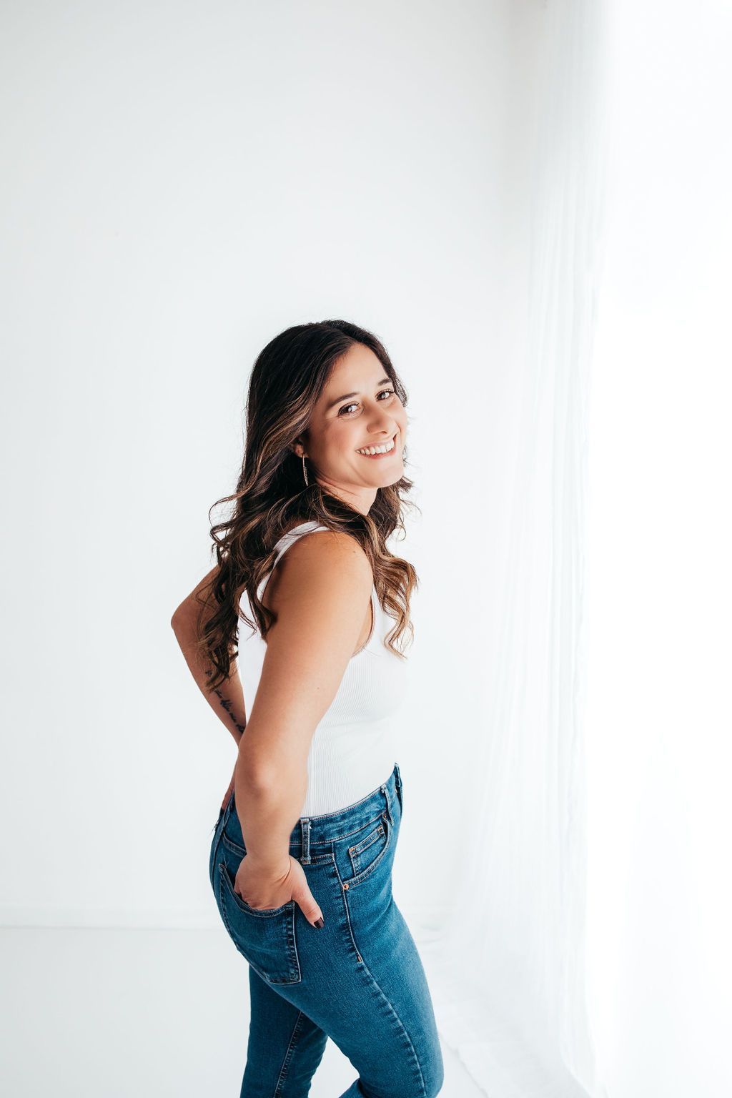 A woman in a white tank top and blue jeans is standing in front of a white wall.