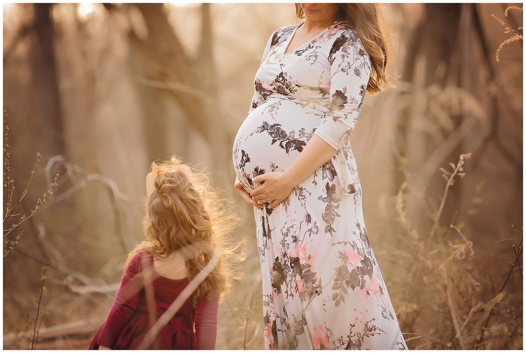 A pregnant woman in a floral dress is standing next to a little girl in a field.