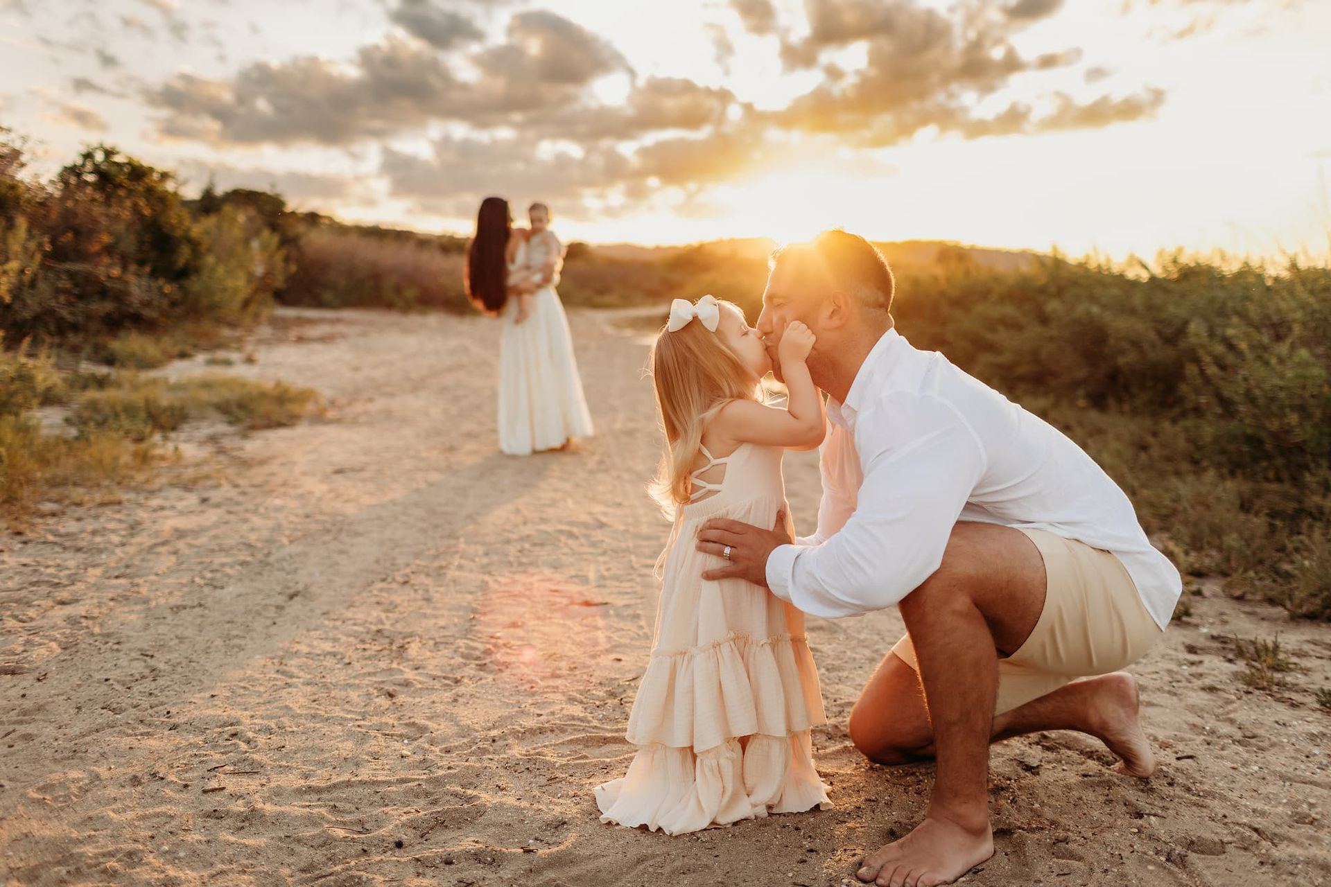 Father kisses daughter on dirt path, mother holds baby in the distance, sunset.