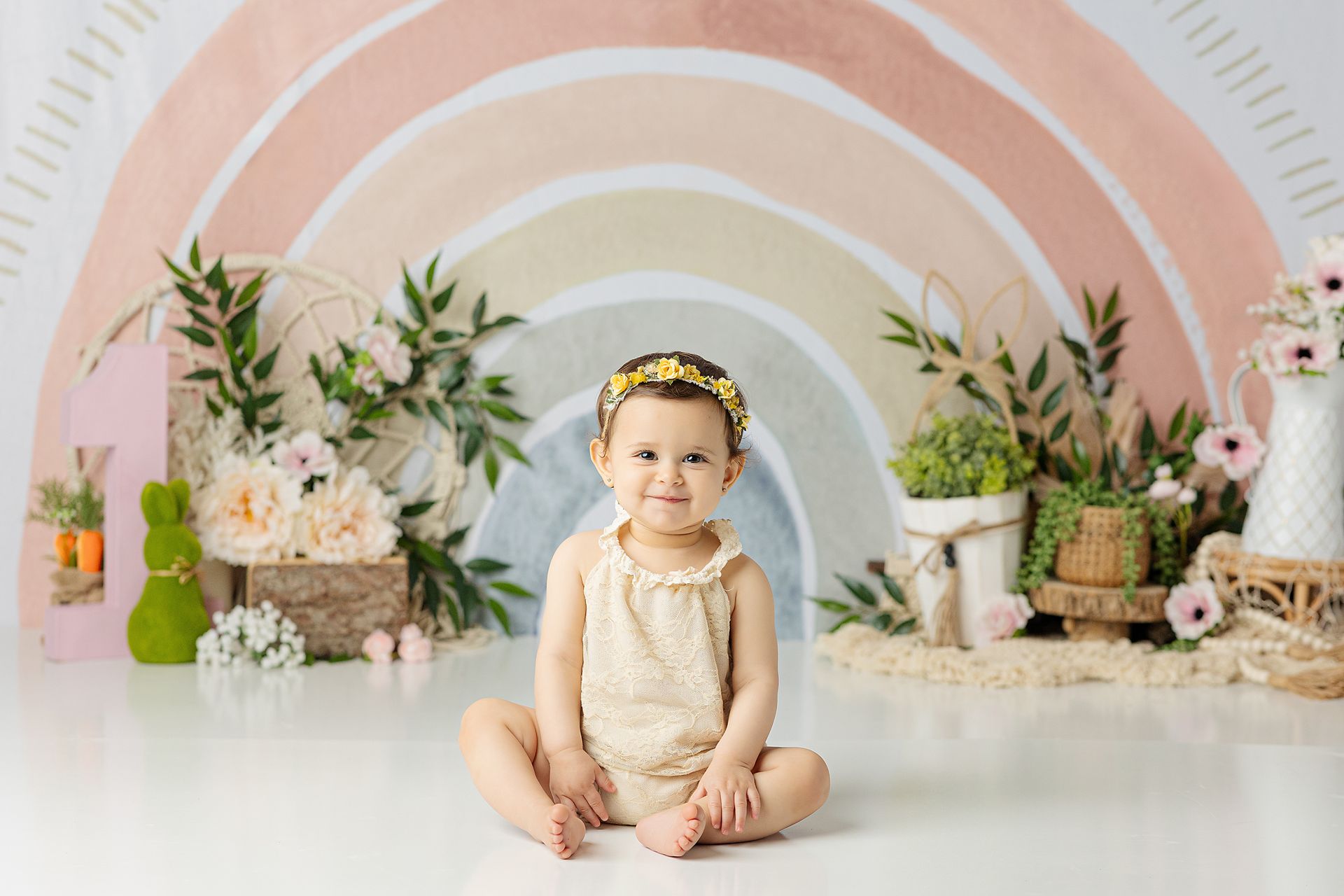 A smiling baby sits in front of a pastel rainbow backdrop. They wear a floral headband and a light-colored onesie.