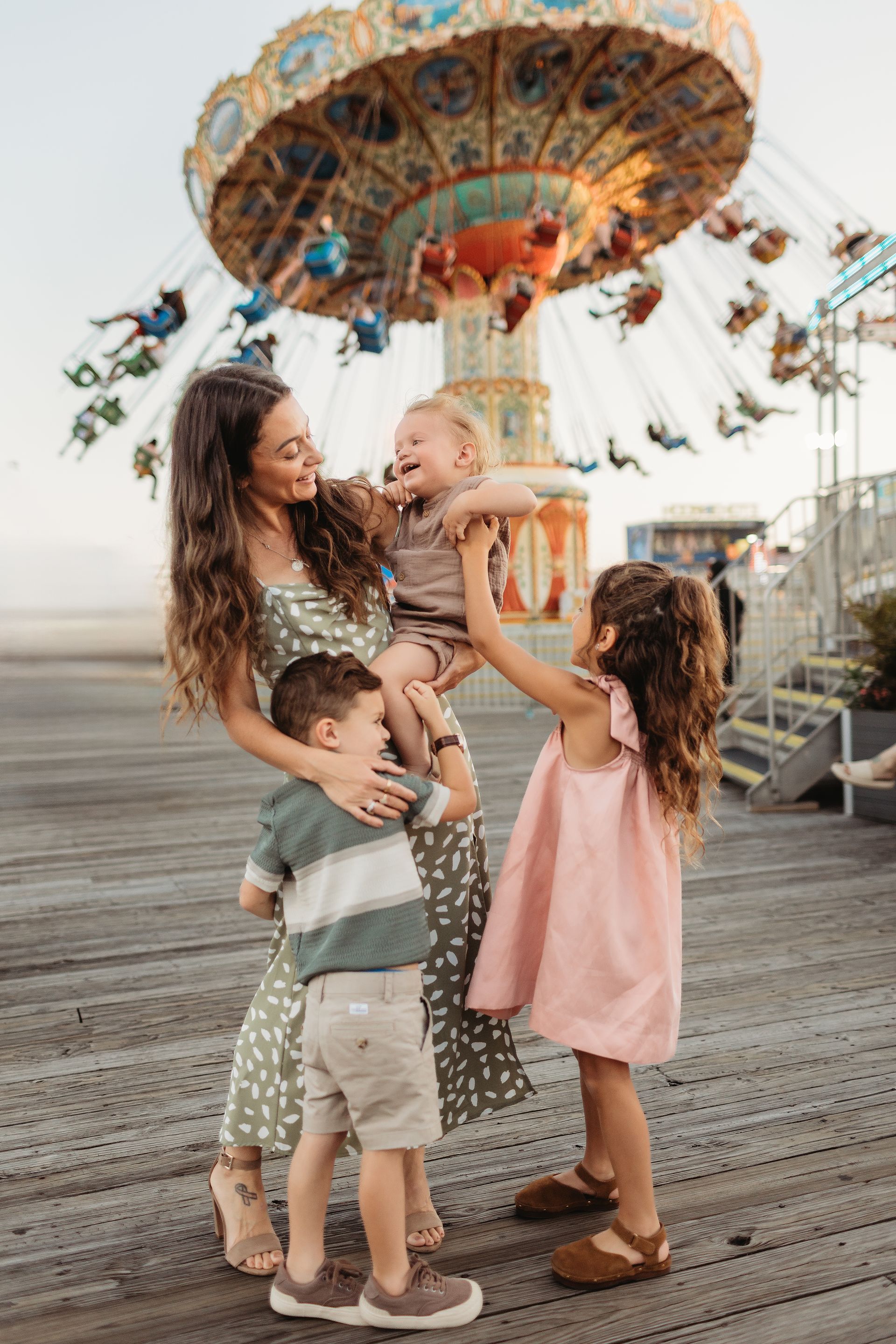 Woman and three children at amusement park; woman holding laughing child, others smiling, carousel in background.