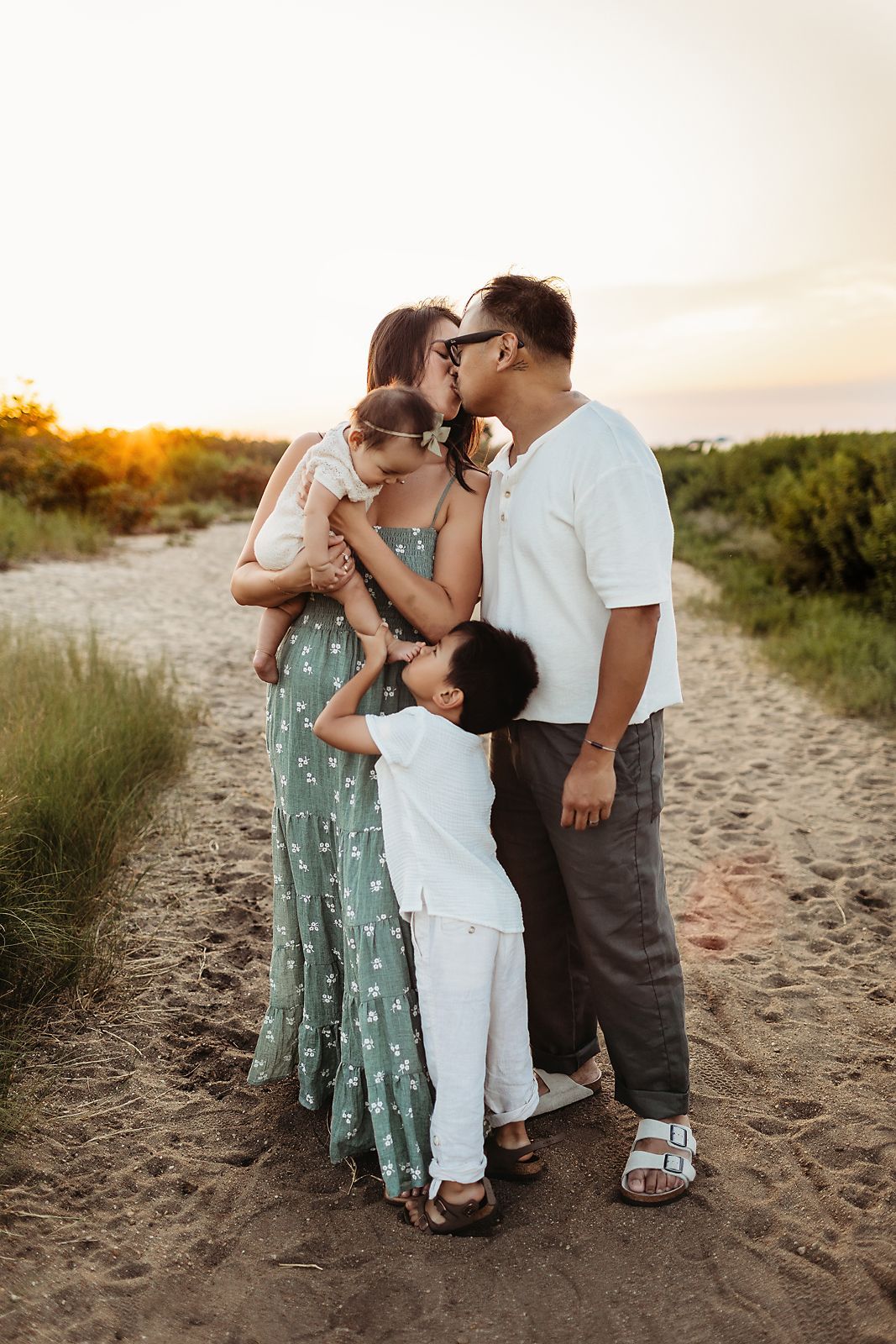 Family of four on a sandy path, parents kissing, holding a baby, with a child hugging his mother's leg at sunset.