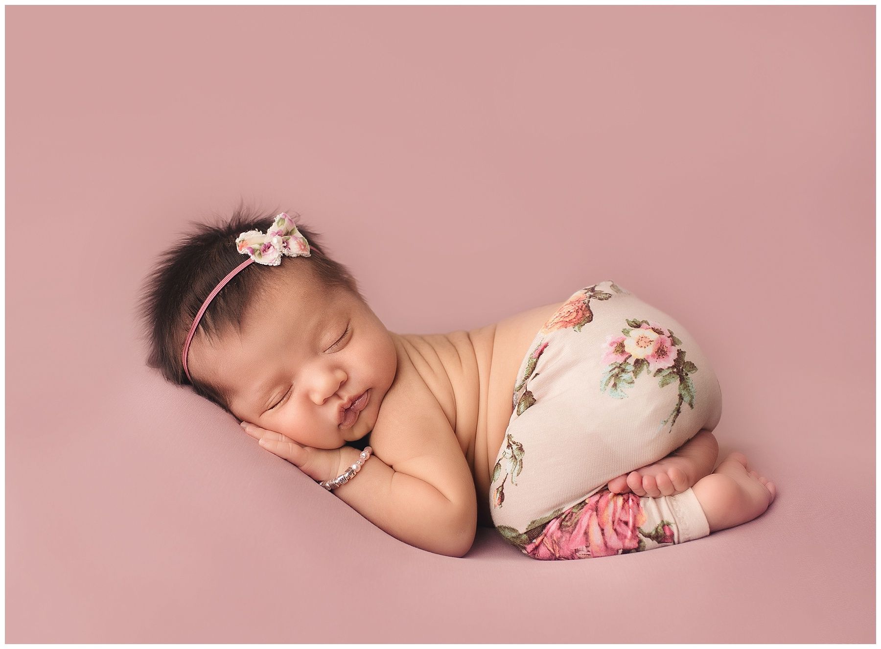 A newborn baby girl is sleeping on her stomach on a pink background.