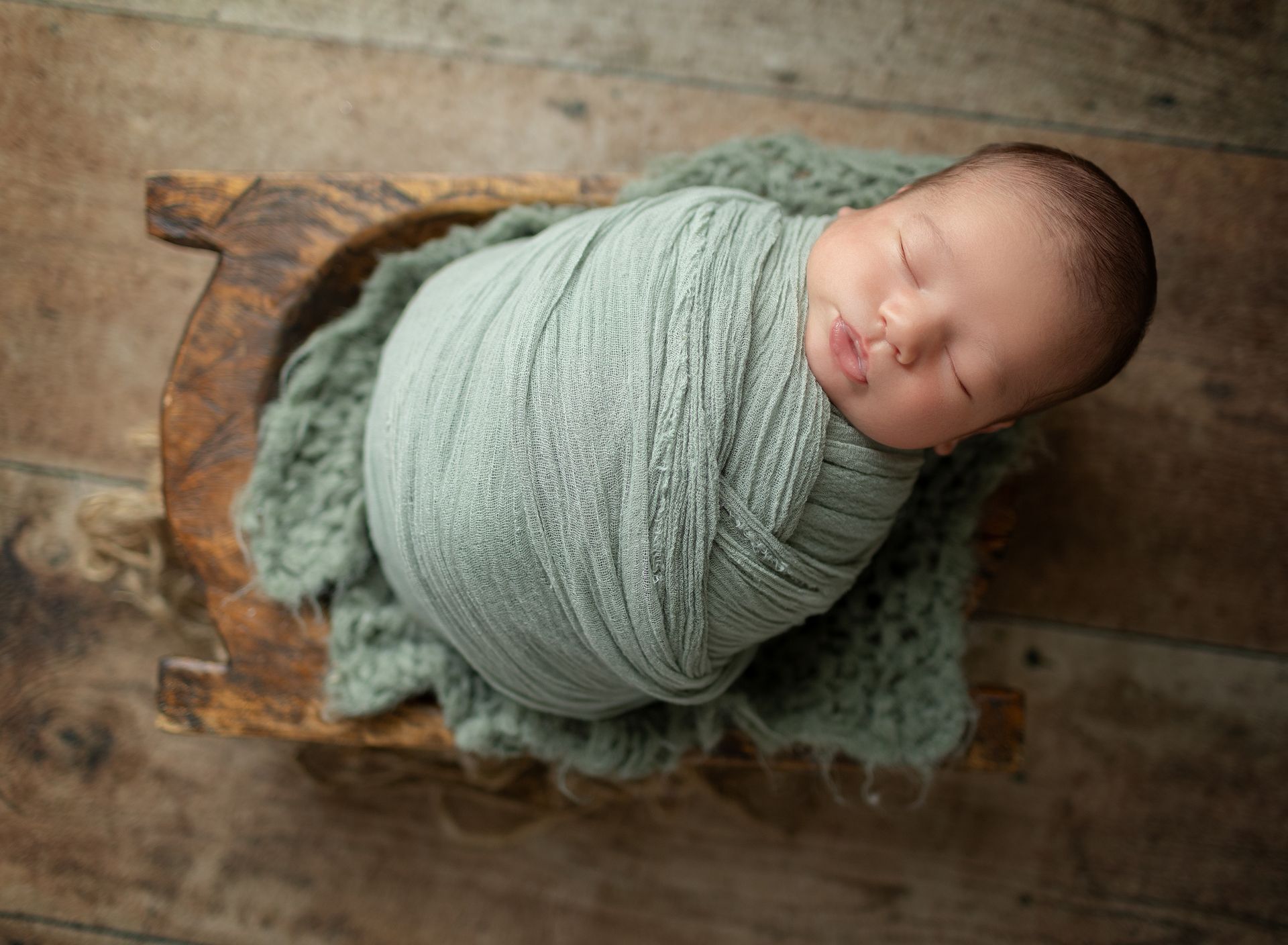 A newborn baby wrapped in a green blanket is sleeping on a wooden sleigh.