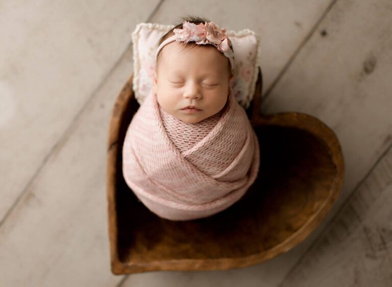 A newborn baby wrapped in a pink blanket is sleeping in a heart shaped wooden bowl.