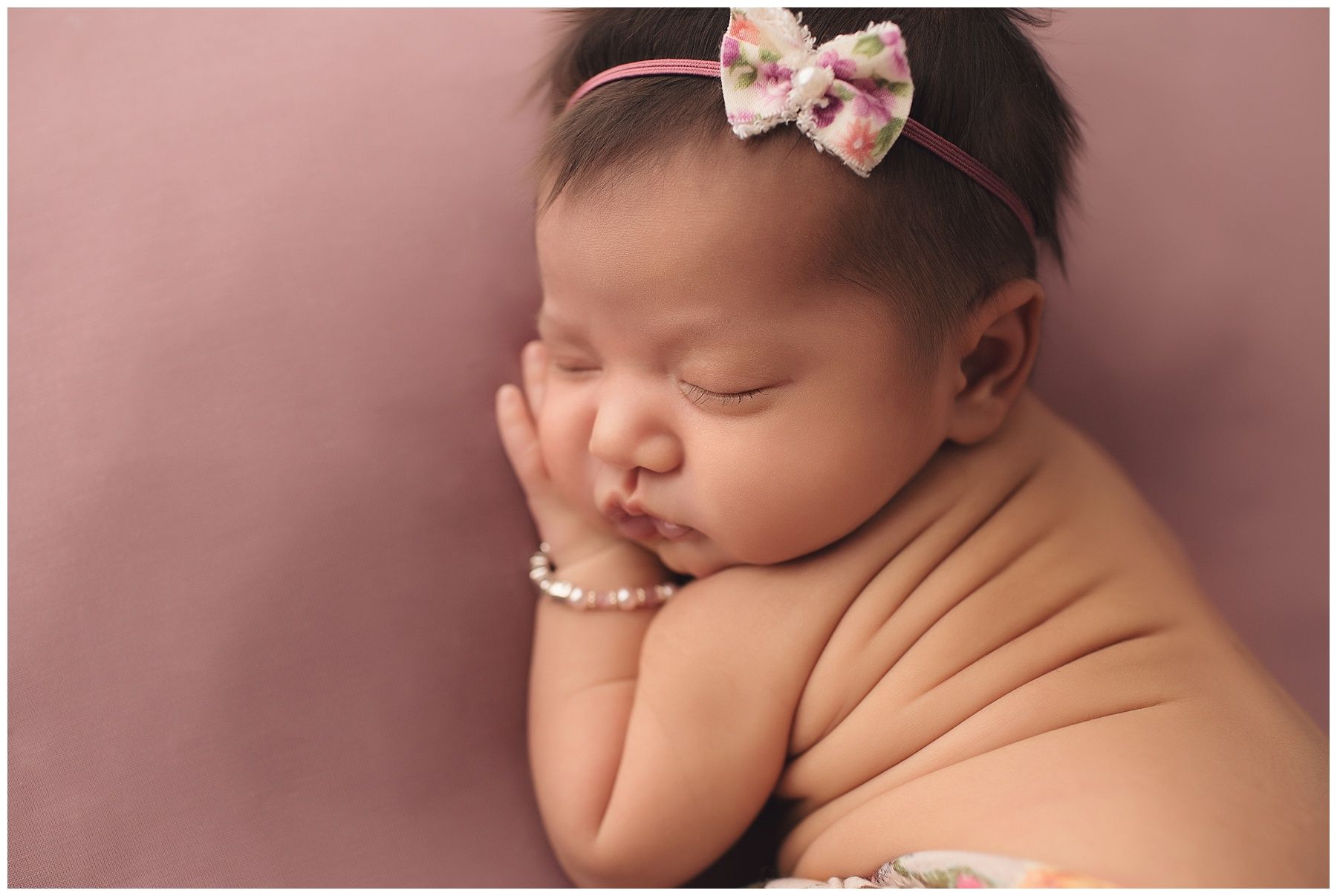 A newborn baby girl wearing a headband and bracelet is sleeping on a pink blanket.