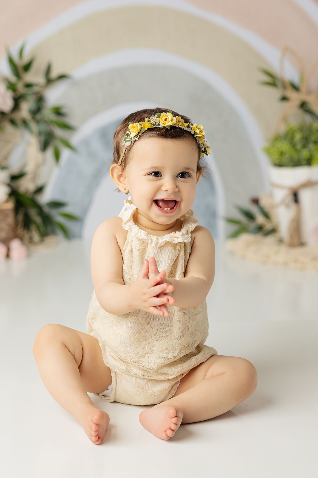 Smiling baby with yellow floral crown, clapping hands, wearing cream lace romper, sitting in front of a rainbow backdrop.