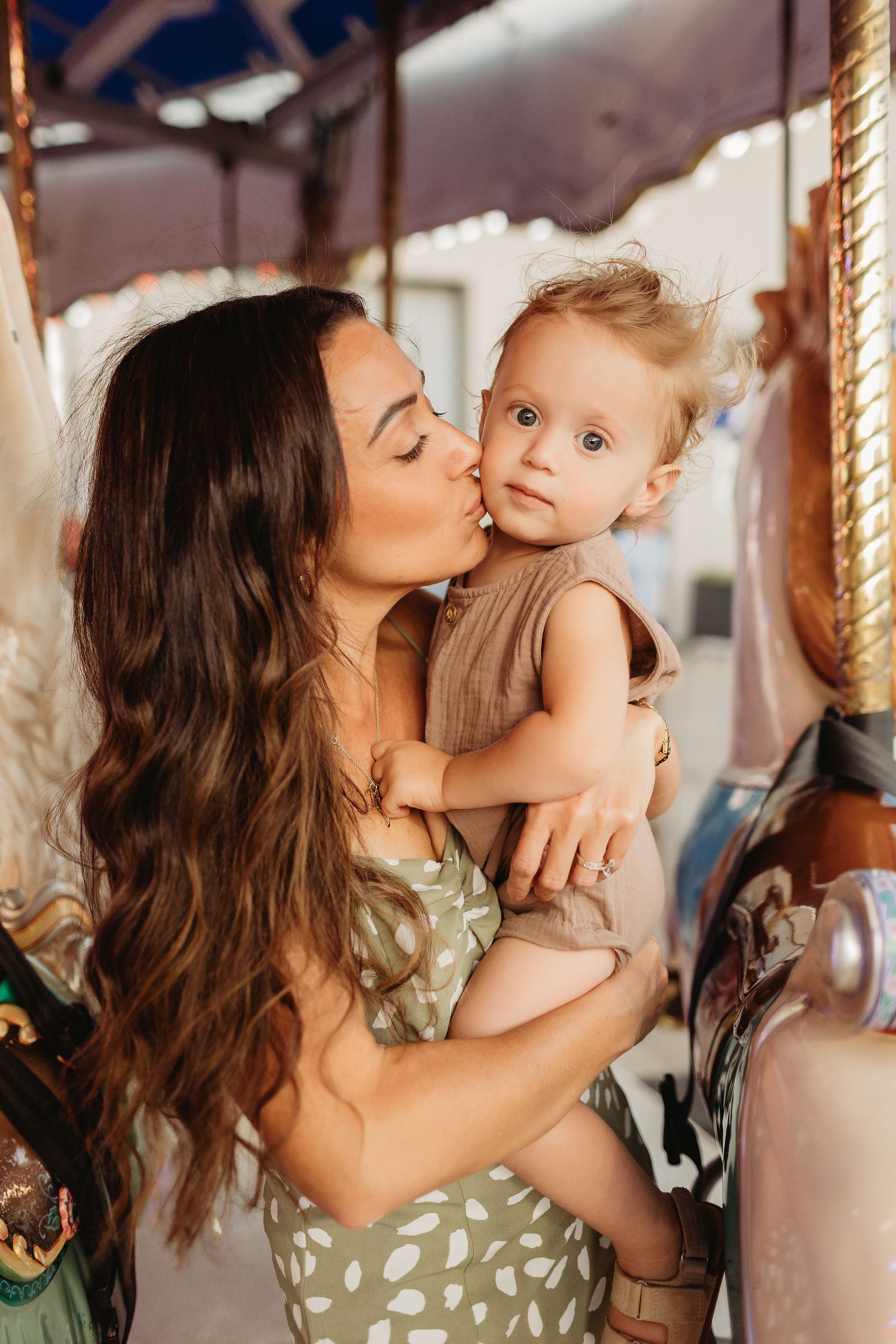 Woman kisses a toddler's cheek at a carousel. They are surrounded by carousel horses.