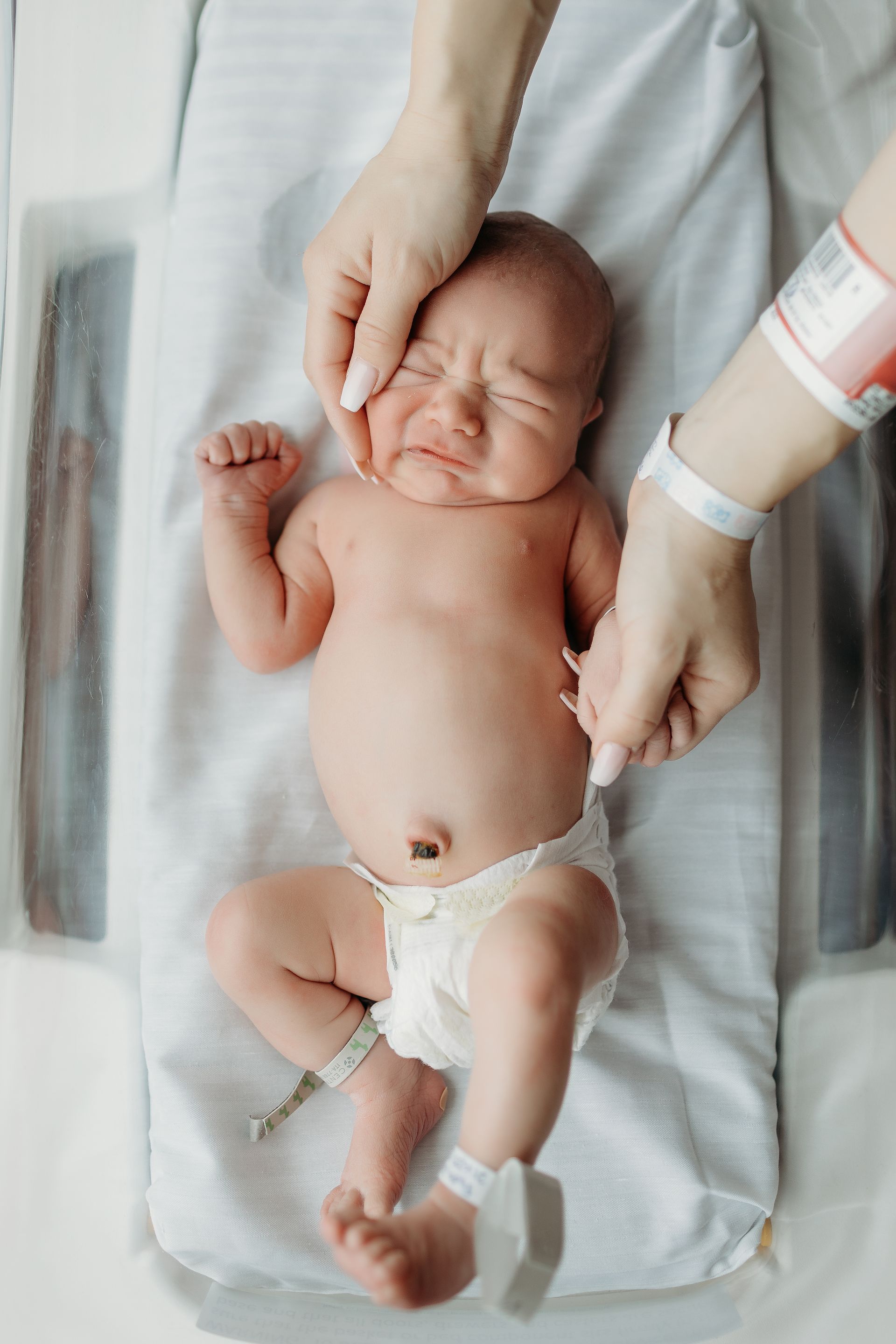 Newborn baby crying on a hospital examination table, being held by two hands.