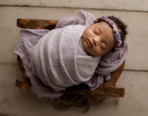 A newborn baby wrapped in a purple blanket is sleeping in a wooden crate.
