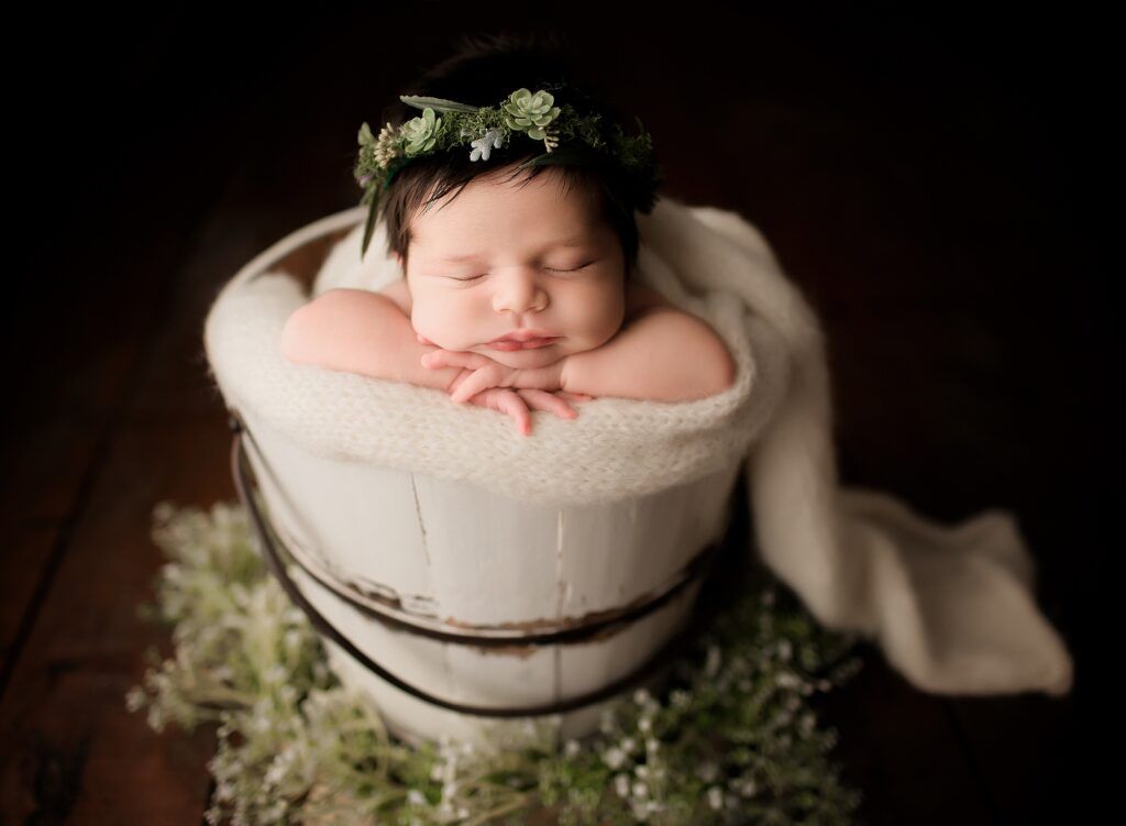 A newborn baby is sleeping in a white bucket with a flower crown on her head.