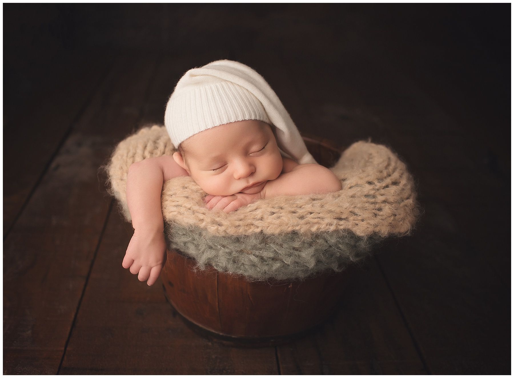 A newborn baby wearing a white hat is sleeping in a wooden bowl.