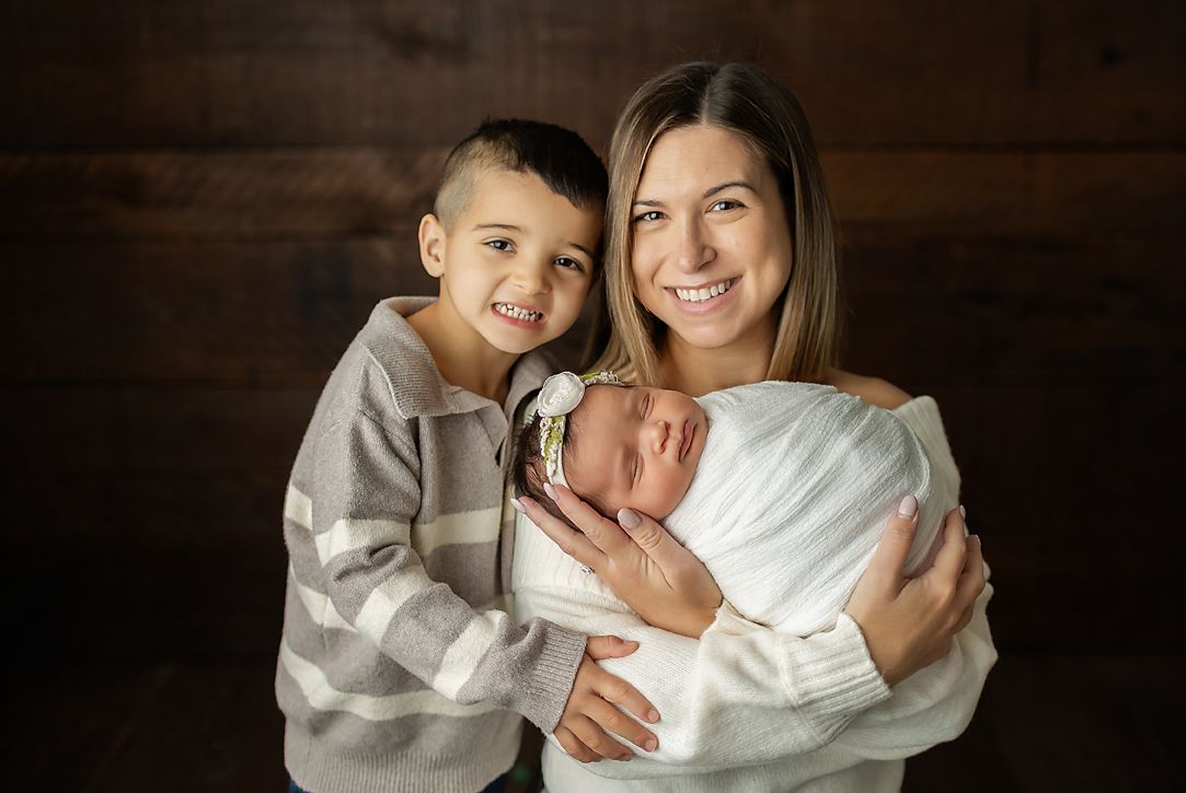 Woman holding a newborn baby, smiles with a young boy next to them. All three in front of a dark wood backdrop.