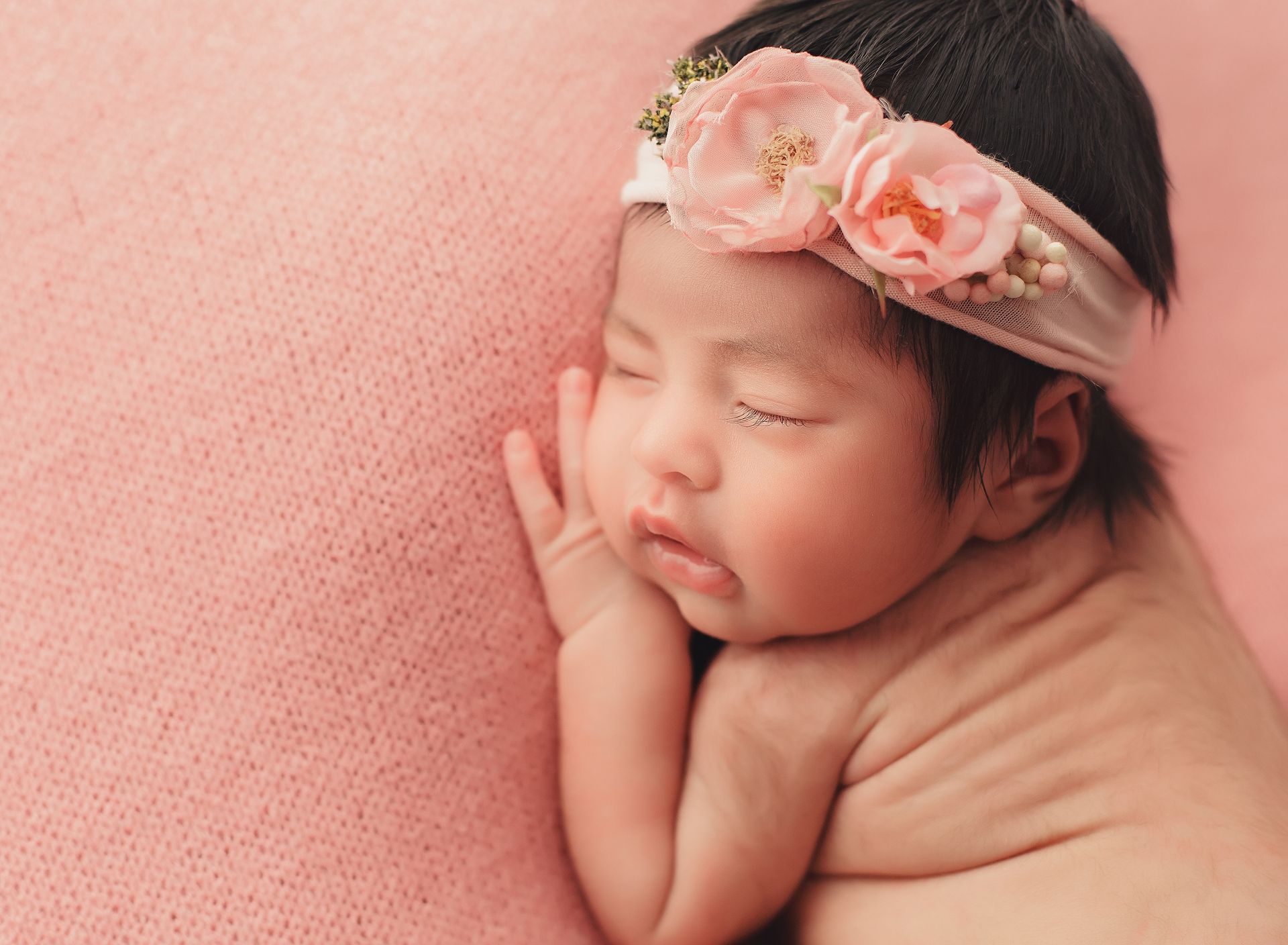 A newborn baby girl wearing a pink headband is sleeping on a pink blanket.