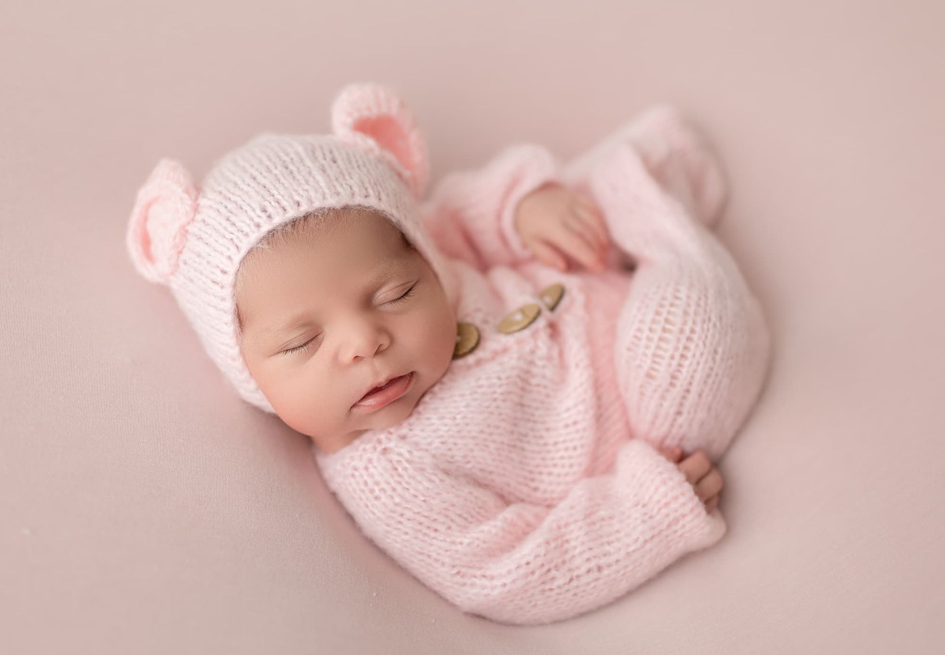 Newborn in pink knitted outfit and hat with ear detail, curled up on pink fabric.