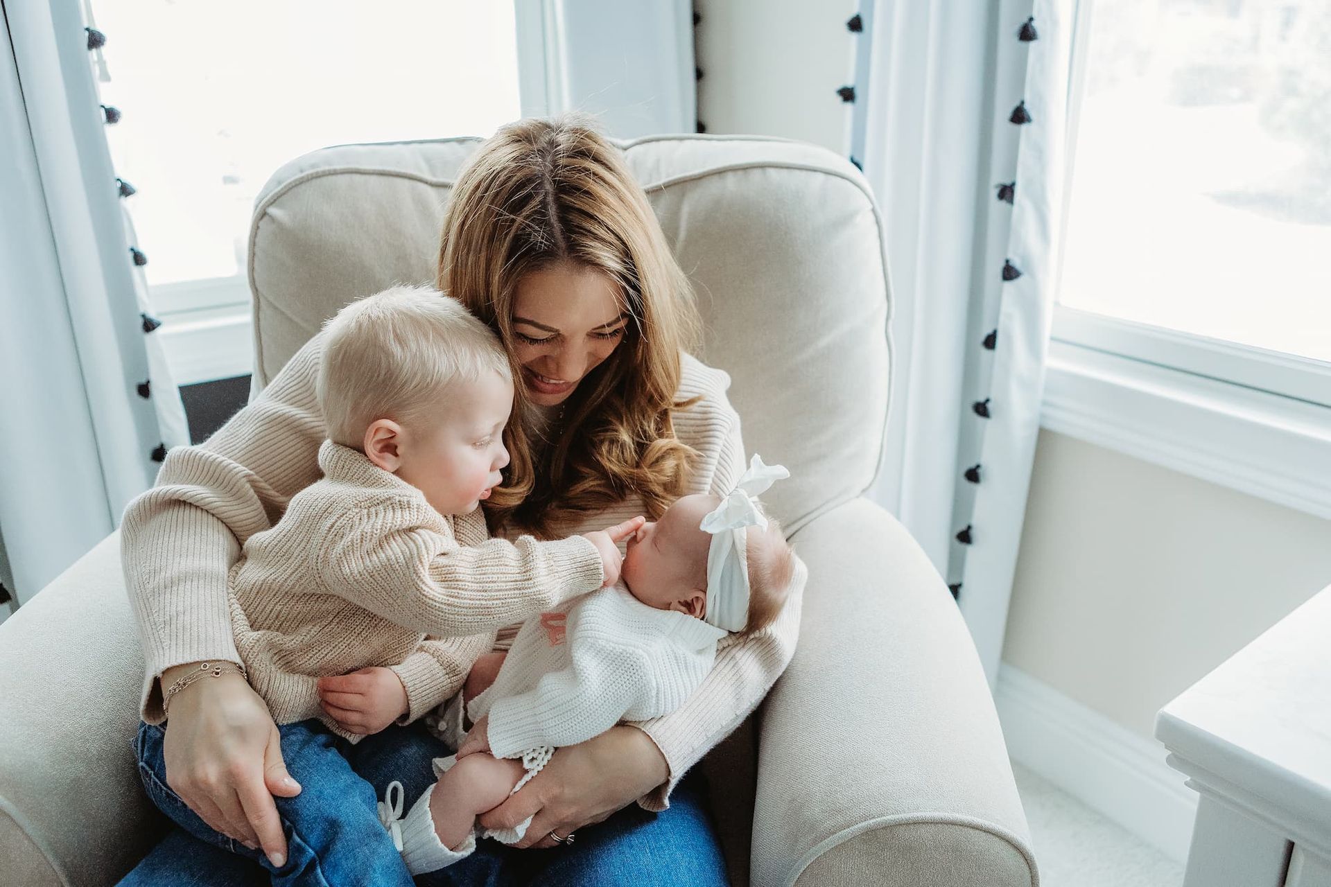 A parent sitting in an armchair holds a baby while a toddler touches the baby’s face, both wearing light-colored sweaters.