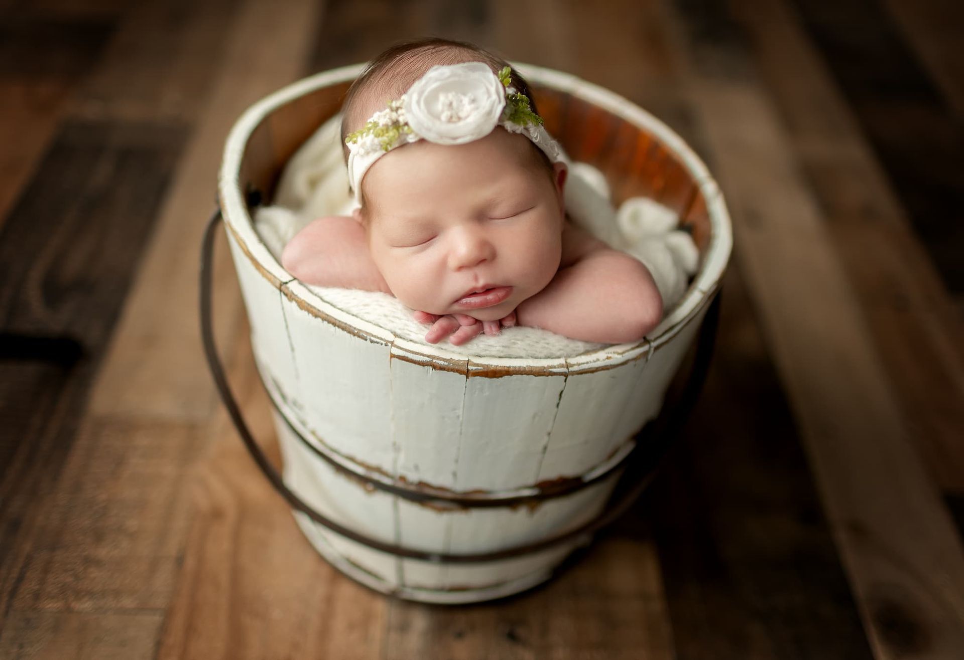 Newborn baby in a white bucket, wearing a floral headband, resting on arms, wooden floor.
