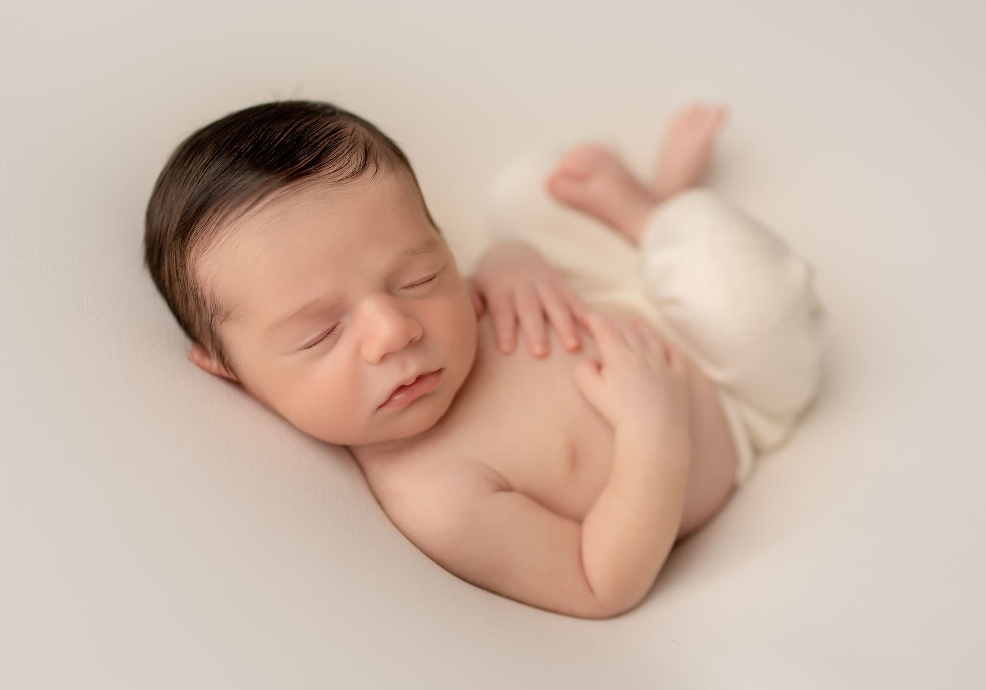 Newborn baby sleeping on a white surface, with dark hair, wrapped in a cream fabric.