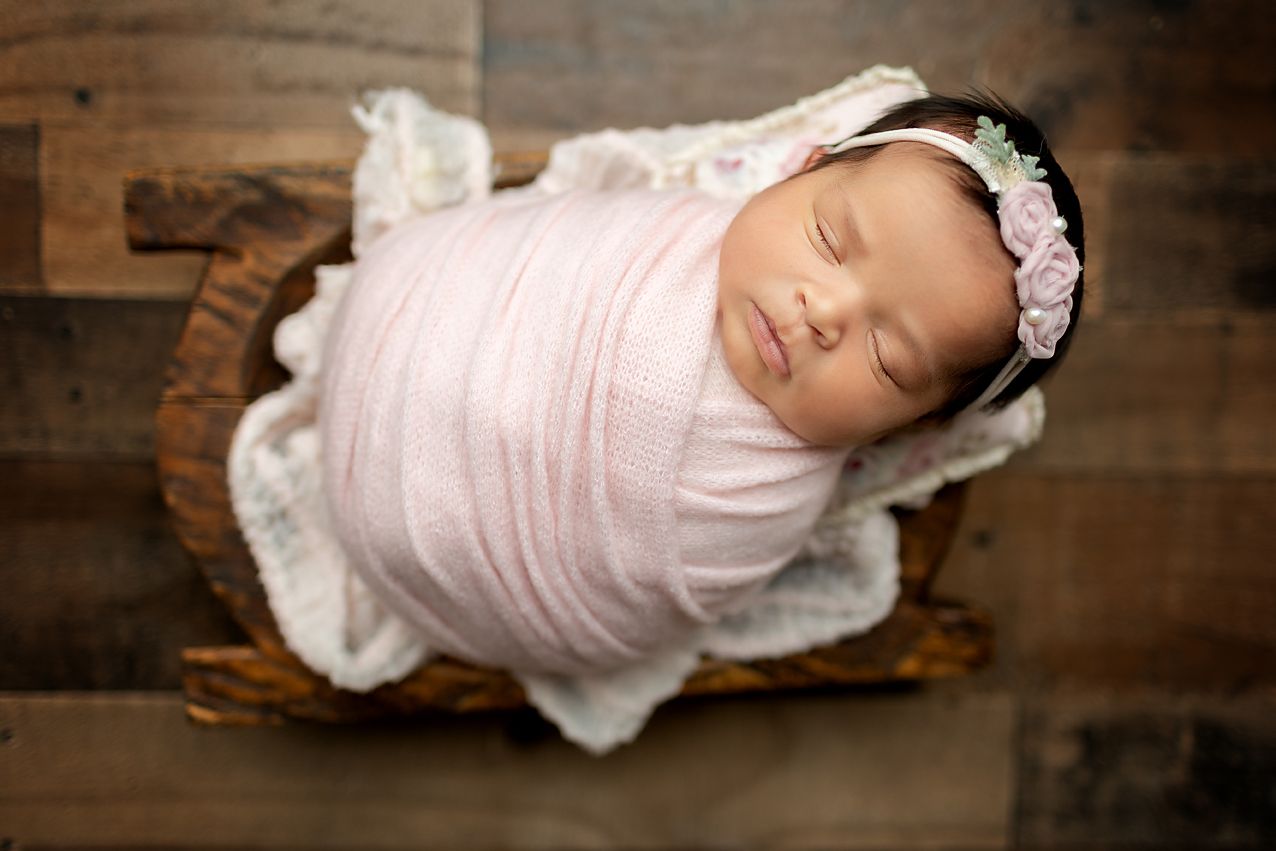 Newborn wrapped in pink blanket, asleep in a wooden bowl, with a floral headband.