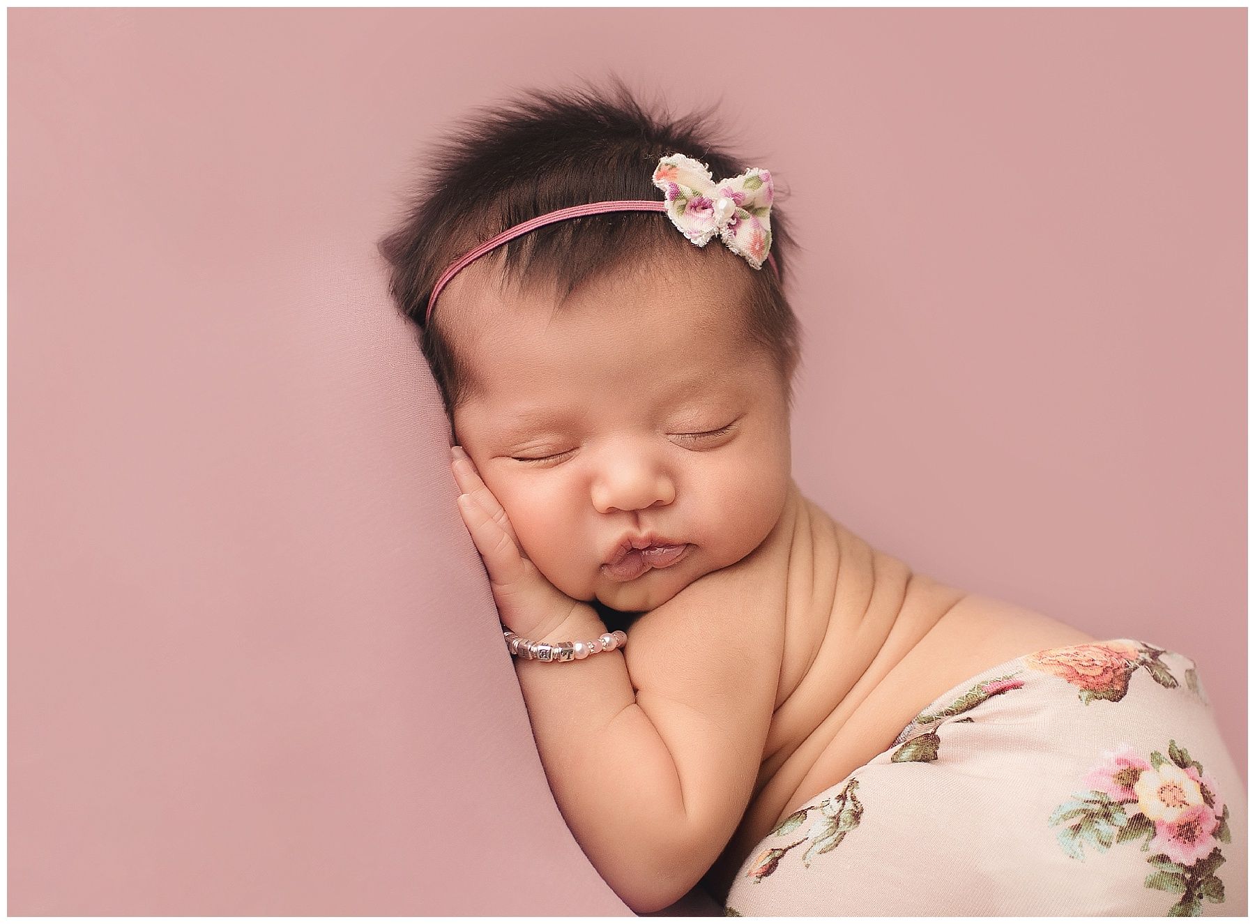 A newborn baby wrapped in a floral blanket is sleeping with her hand on her face.