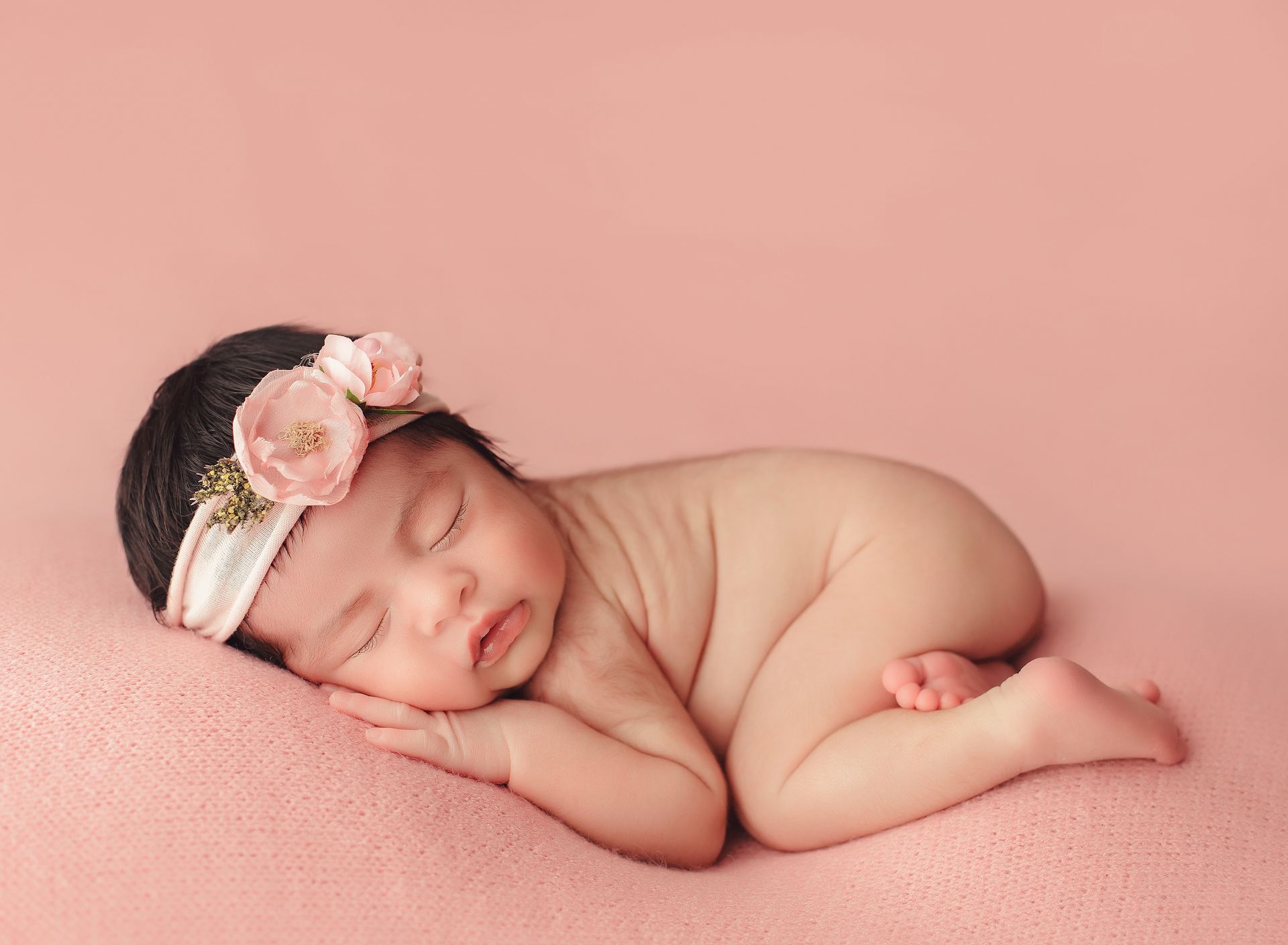 A newborn baby girl wearing a headband is sleeping on a pink blanket.