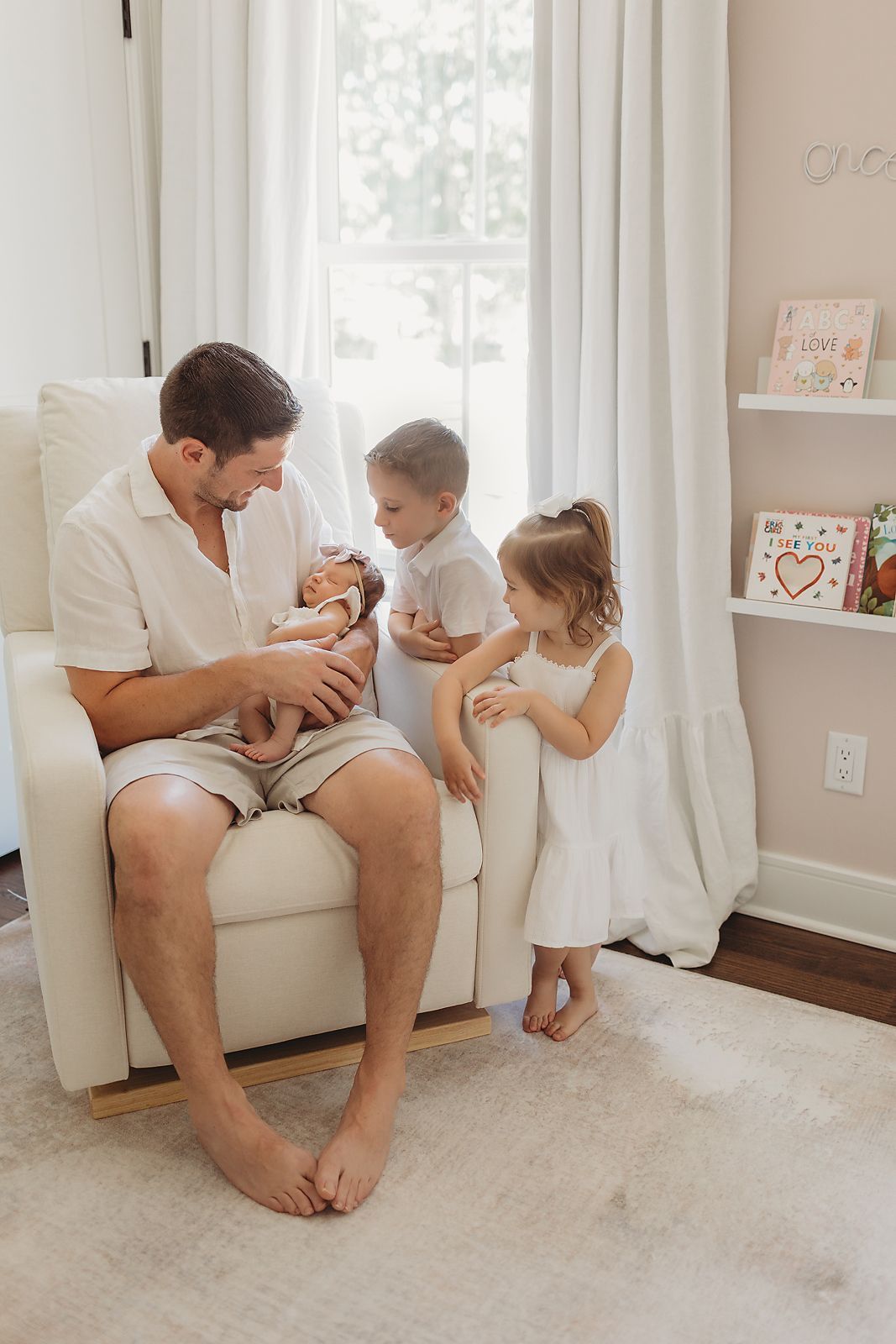 Father sitting in chair holding newborn, two children looking on. Nursery setting, neutral colors.