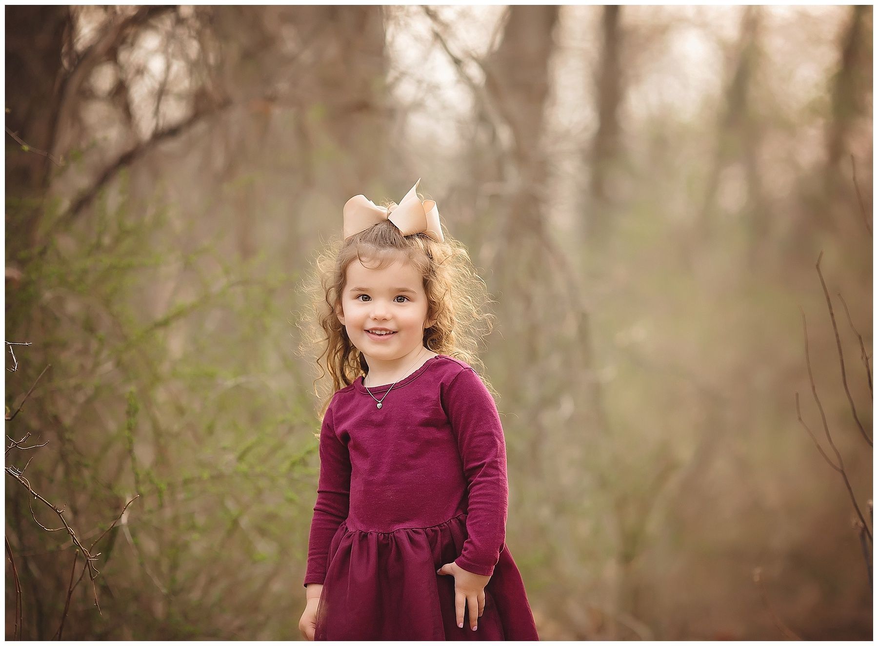 A little girl in a purple dress with a bow on her head is standing in the woods.