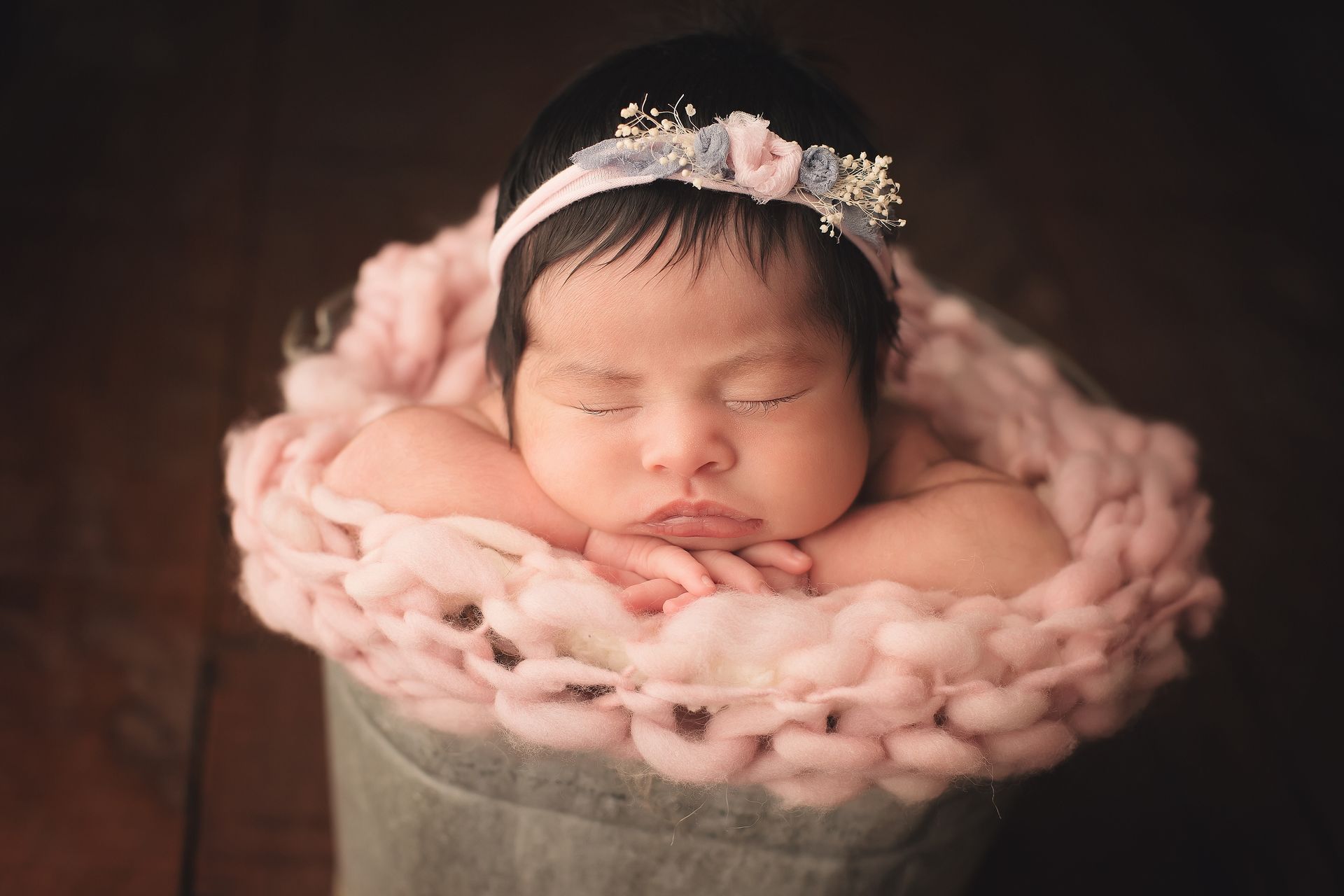A newborn baby girl is sleeping in a bucket with a pink headband.