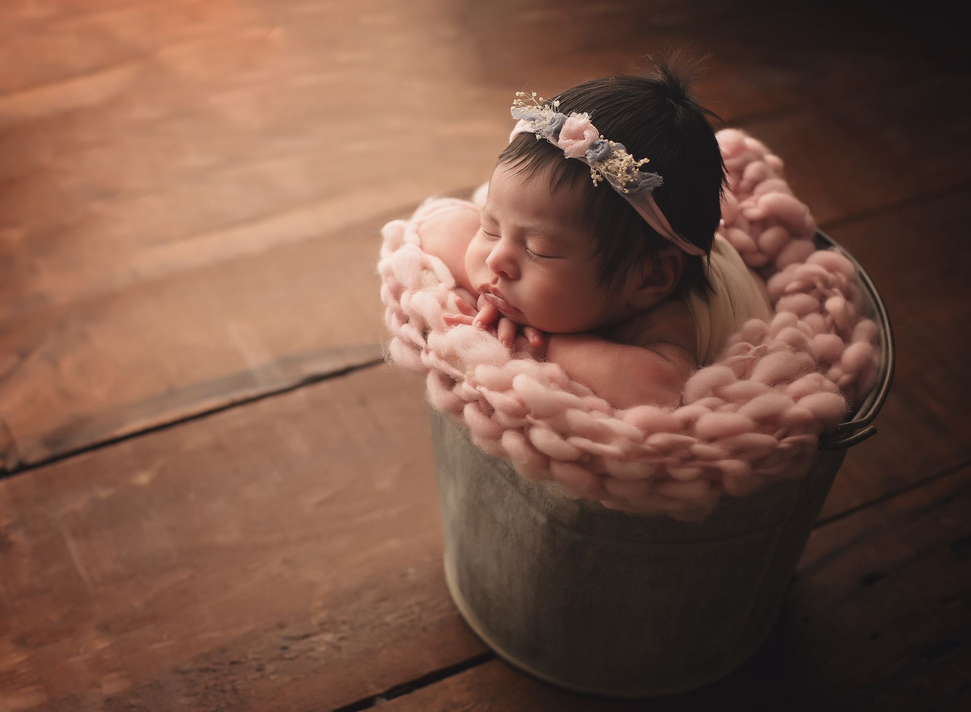 A newborn baby is sleeping in a bucket on a wooden floor.