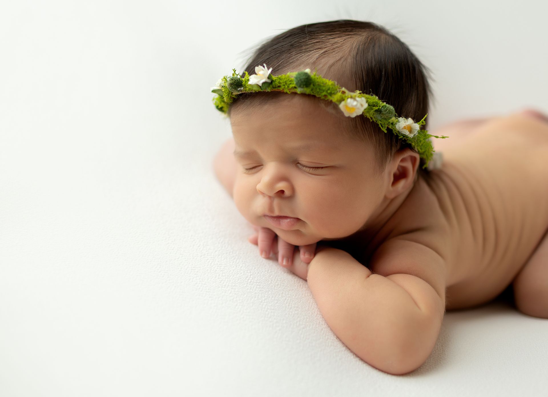 Newborn baby, wearing a floral crown, lying on stomach with hands under chin, eyes closed on white surface in Ridgewood, NJ