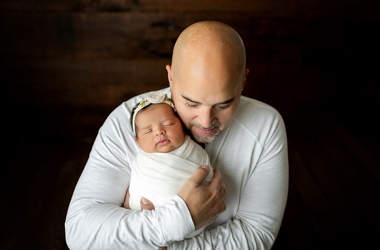 Man holding newborn, smiling, with older child smiling nearby, set against wood backdrop.