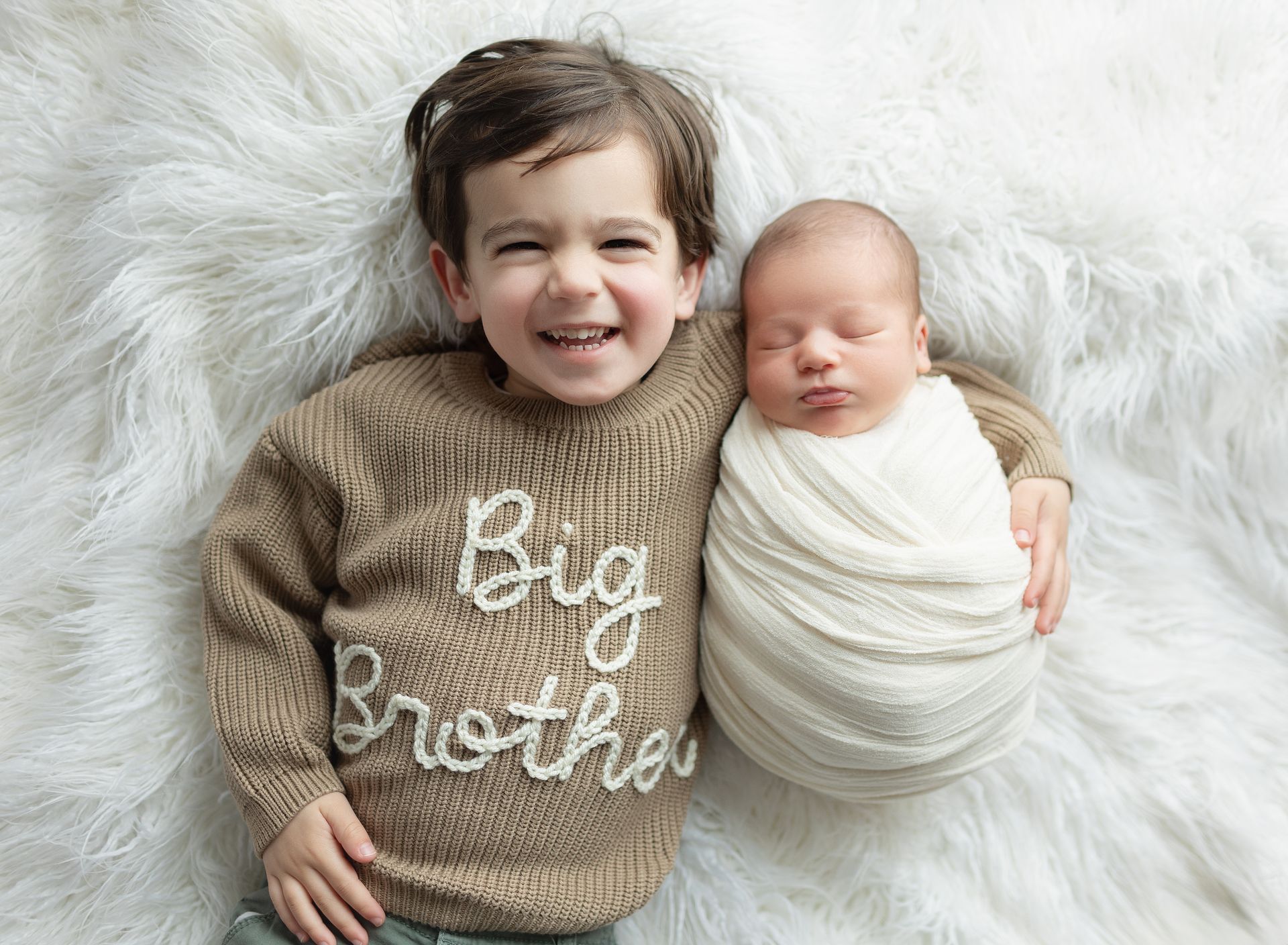 A little boy is laying next to a newborn baby wrapped in a blanket.