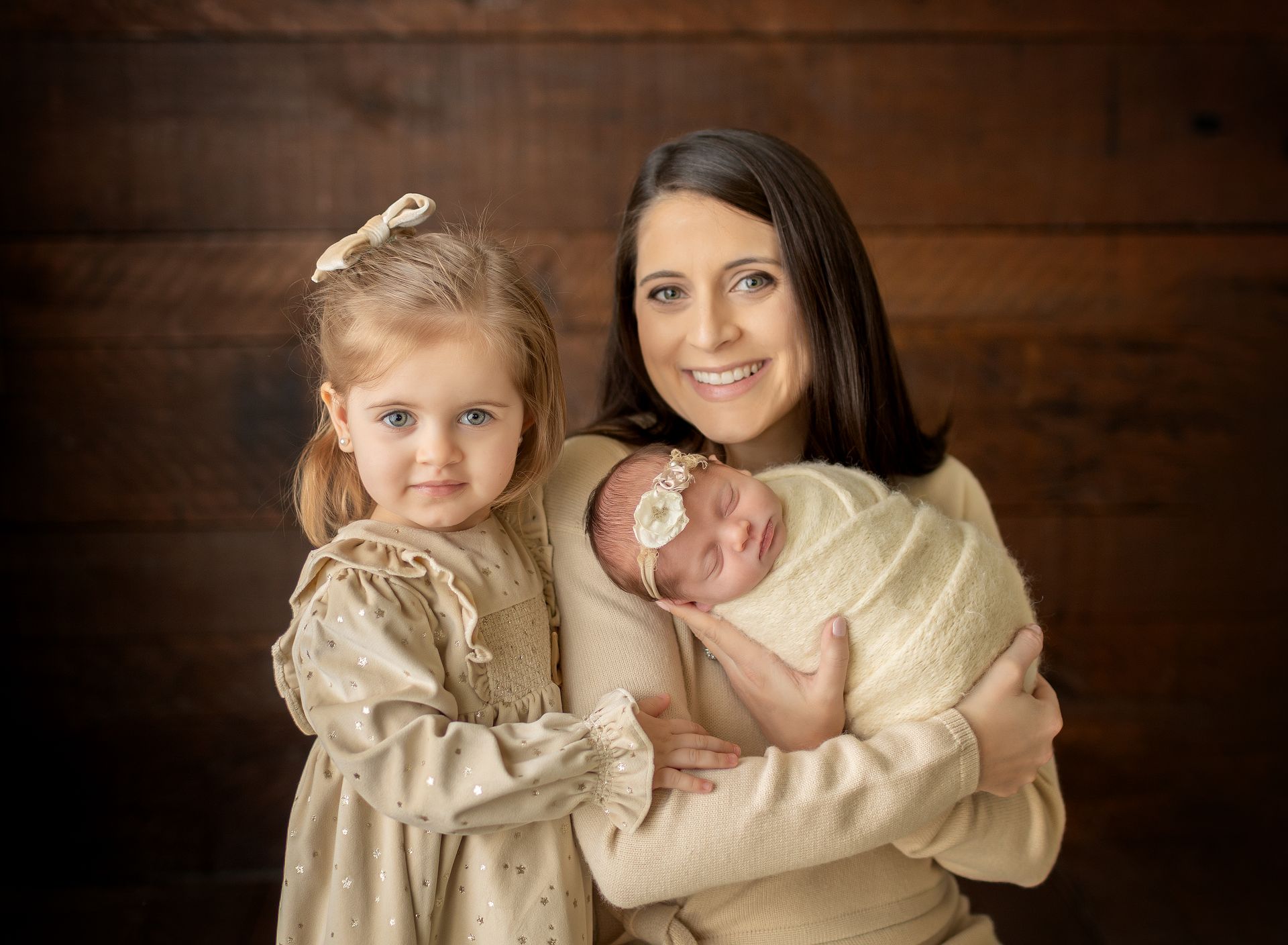 Woman holding newborn, with young child, against a wood background. All are wearing beige in Passaic County, NJ.