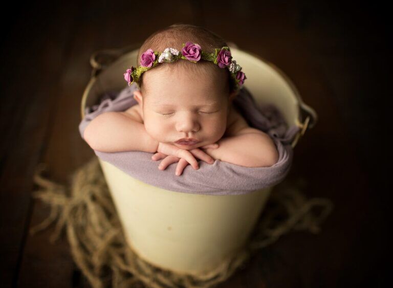 A newborn baby is sleeping in a white bucket with a flower crown on her head.