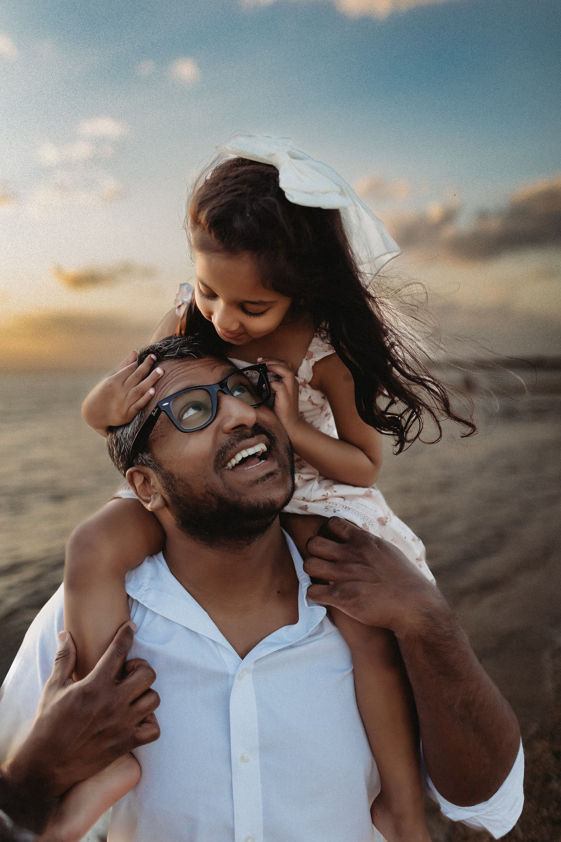 Father smiles while daughter sits on his shoulders, kissing his cheek at the beach.