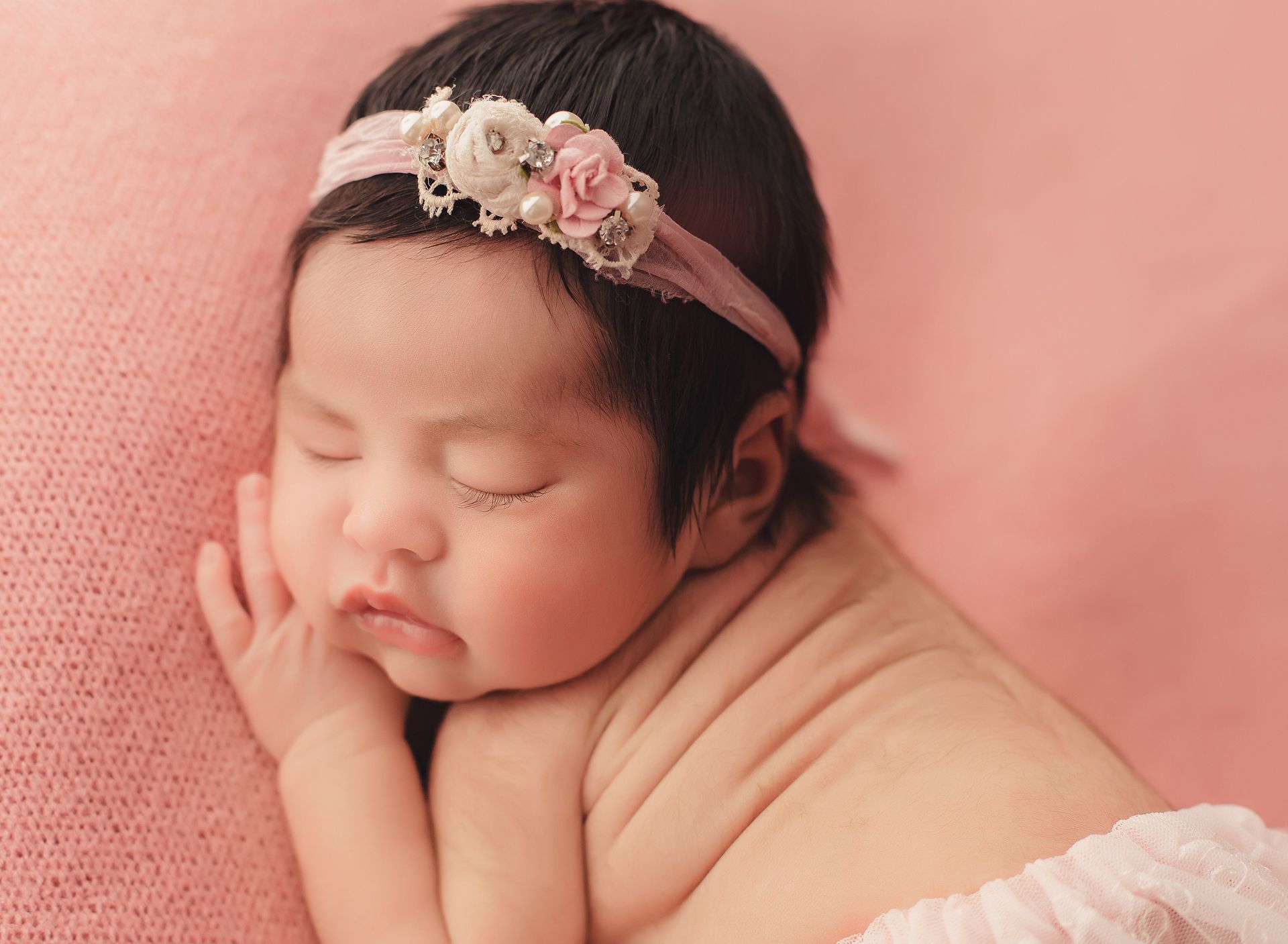 A newborn baby girl wearing a headband is sleeping on a pink blanket.