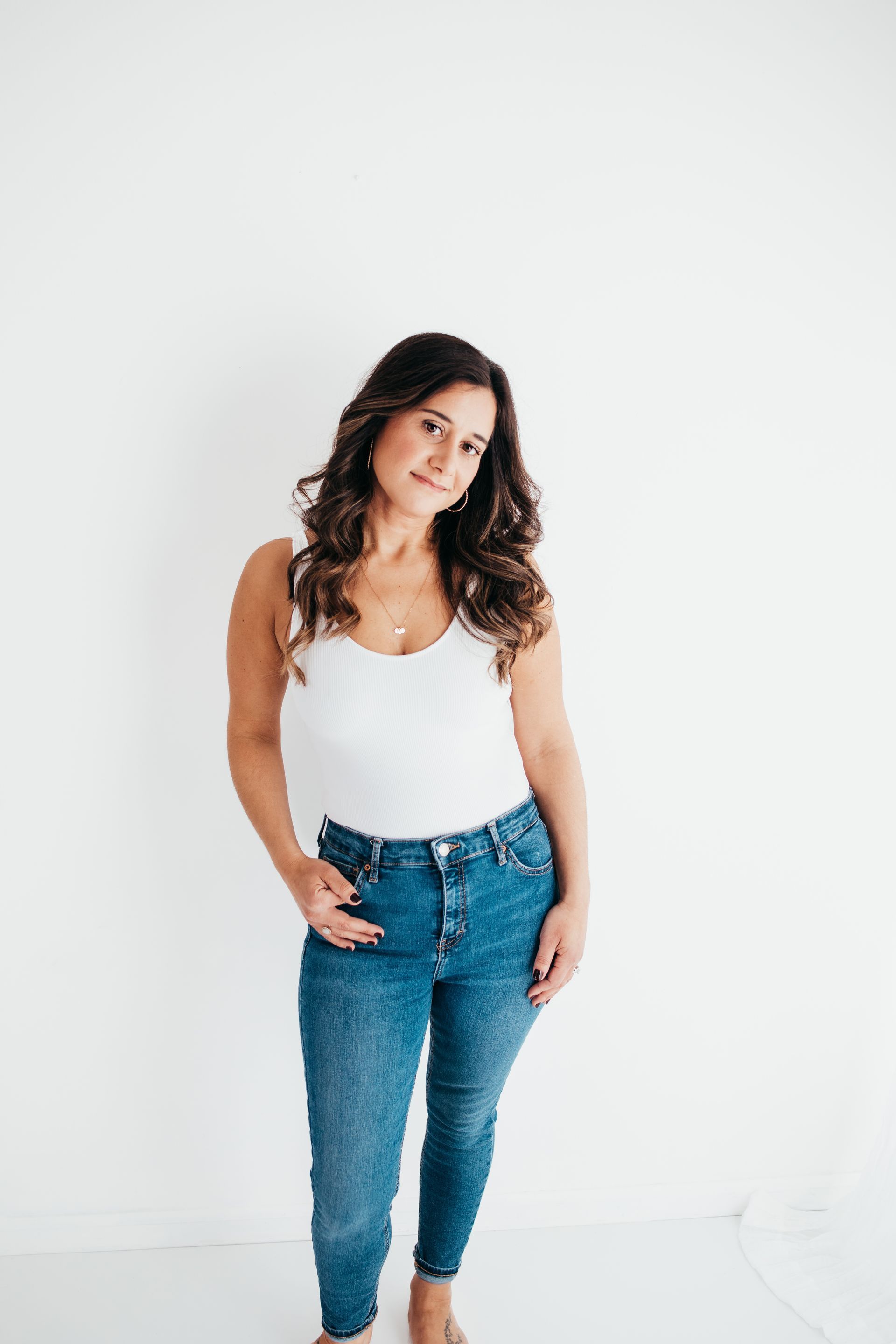 A woman in a white tank top and blue jeans is standing in front of a white wall.