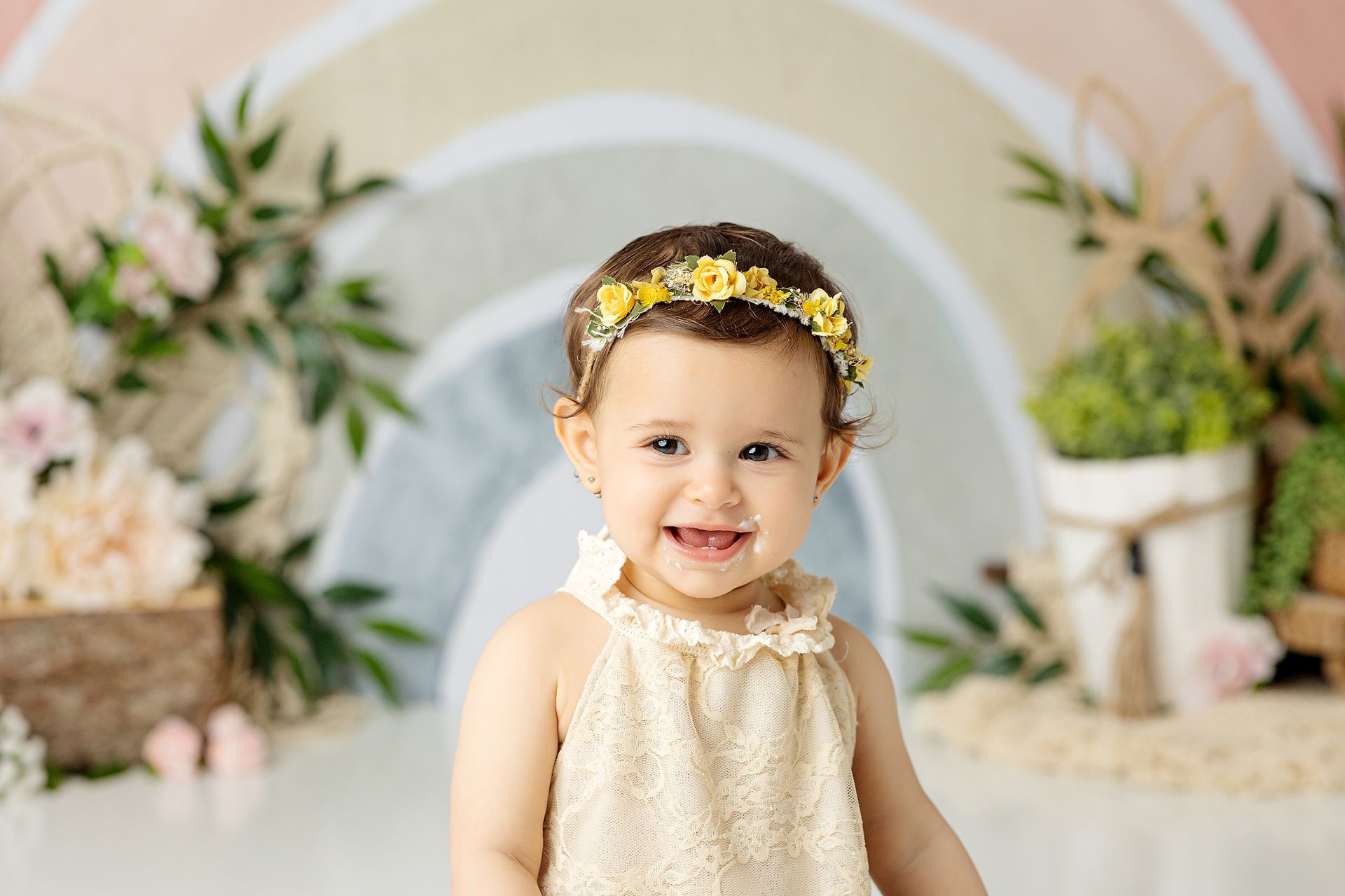 Smiling toddler wearing floral headband, in front of a pastel rainbow backdrop.