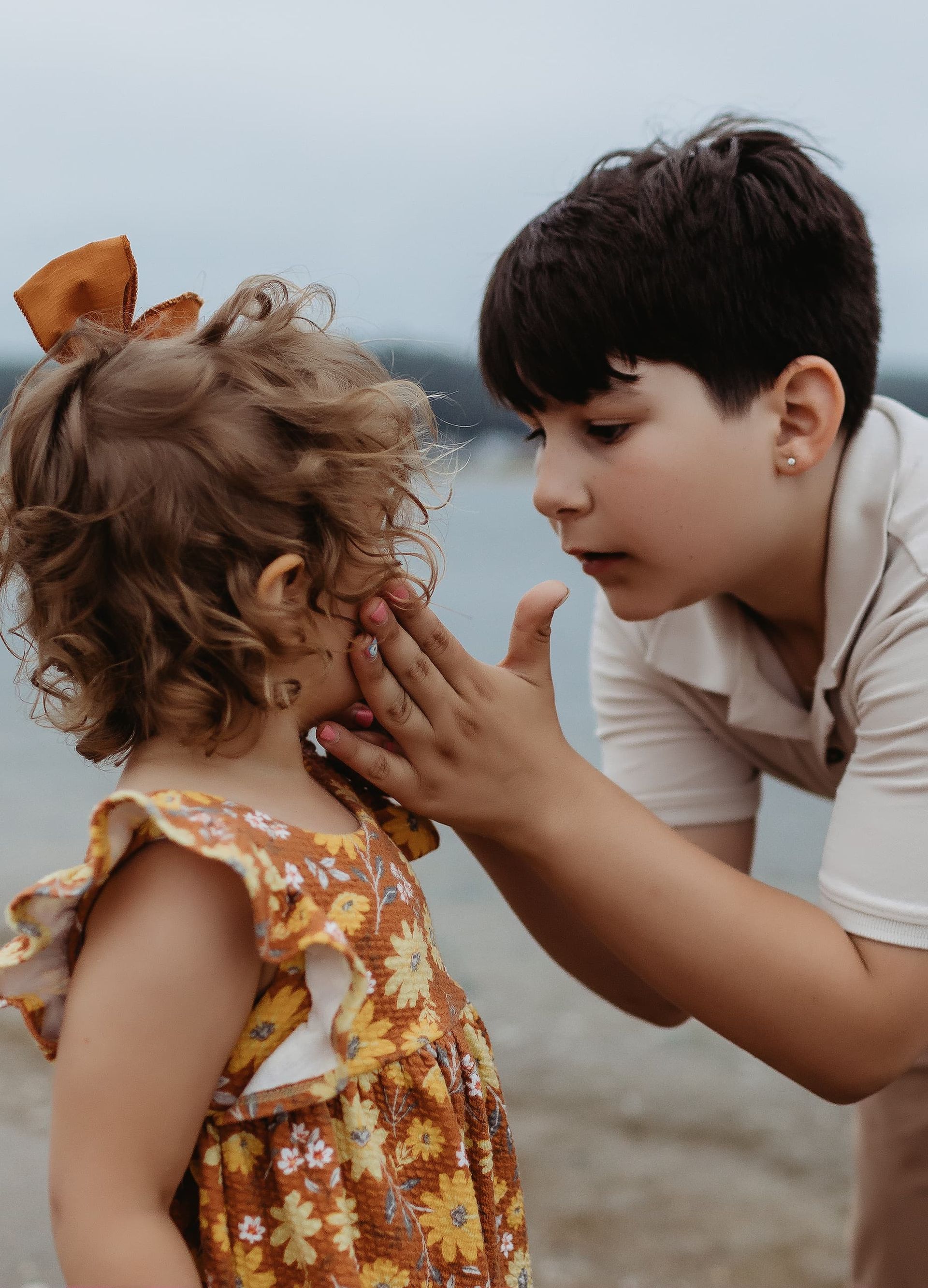 Boy gently touches the cheek of a young child at the beach. Both are looking at each other, the boy concerned.