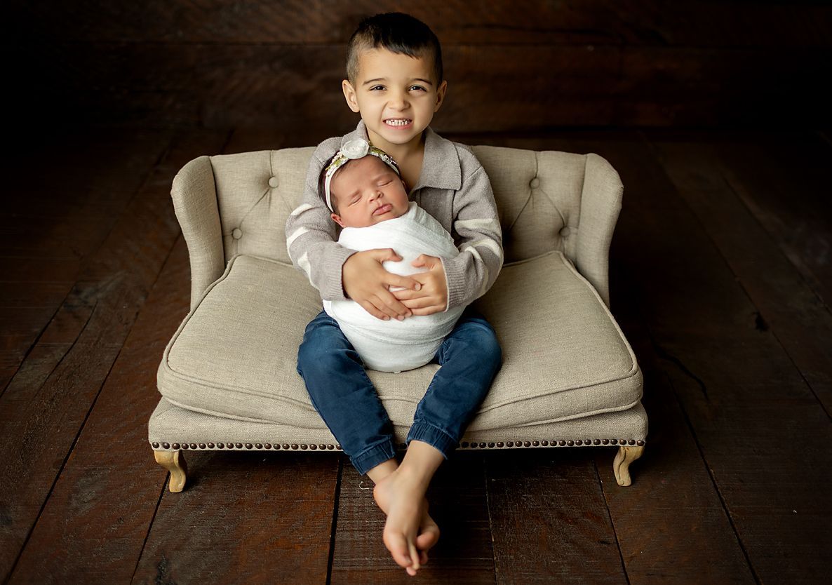 Boy holding a newborn baby wrapped in a blanket, sitting on a small sofa on a wooden floor.